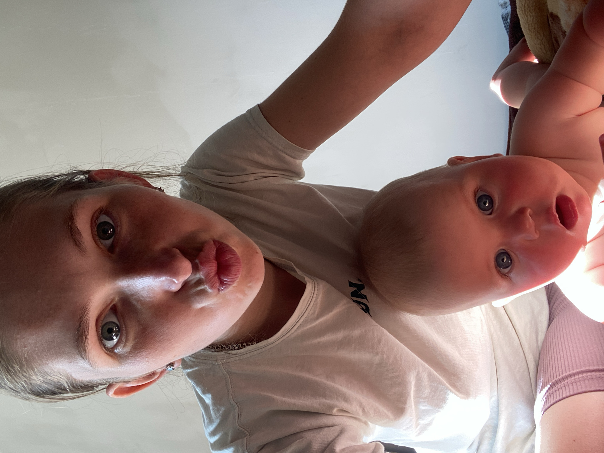 In this upward-facing selfie, a young woman and a baby are the subjects. The woman is in the foreground, looking directly at the camera with her lips pursed. She has blonde hair and is wearing a white t-shirt. Next to her, partially obscured, is a baby with fair skin and blue eyes, also looking towards the camera. The baby appears to be held or supported by the woman, with their arm visible. Both subjects are wearing light-colored clothing, suggesting a casual setting. The background is a plain, light-colored surface, possibly a wall or ceiling, with a bright light source casting some glare. The image has a warm, bright feel, potentially indicating daytime. There is no visible text in the image.