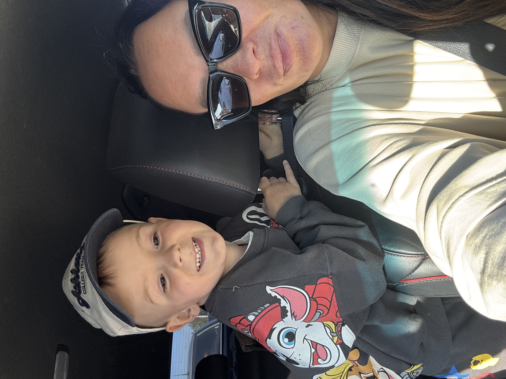 A woman wearing sunglasses and a light-colored sweater, and a young boy wearing a hat and a Paw Patrol sweatshirt, are positioned close together, likely in a car. The boy is in the foreground with the woman behind him. The car has black leather seats. The location is likely Chaikovsky, Russia.