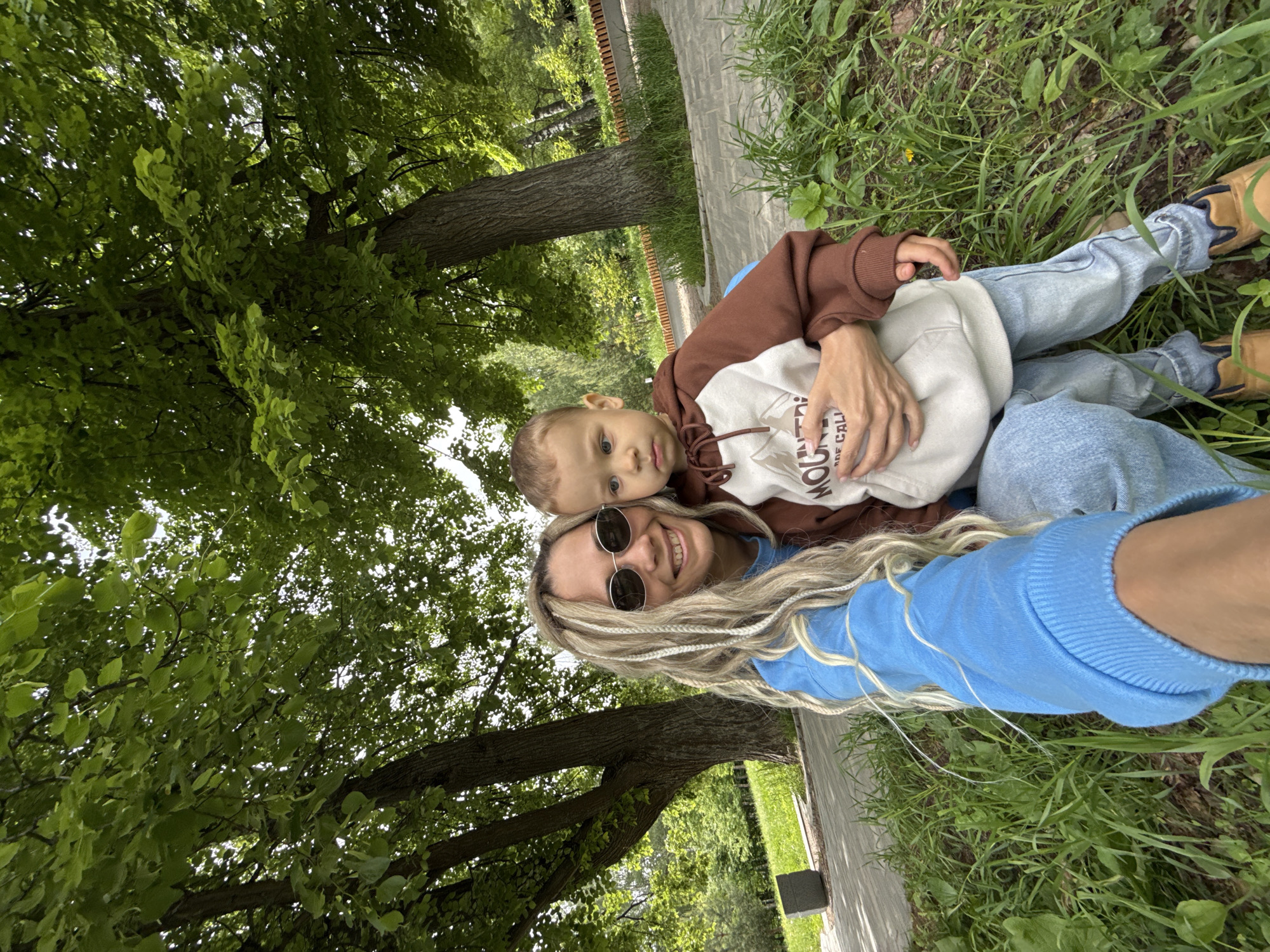 A woman and a child are lying in the grass near a sidewalk in Шебунино, Russia. The woman is wearing a blue sweater and sunglasses, and has long, blonde hair. The child is wearing a brown and white hoodie. They appear to be taking a selfie, with a large tree visible in the background. The grass is green and there is a sidewalk nearby.