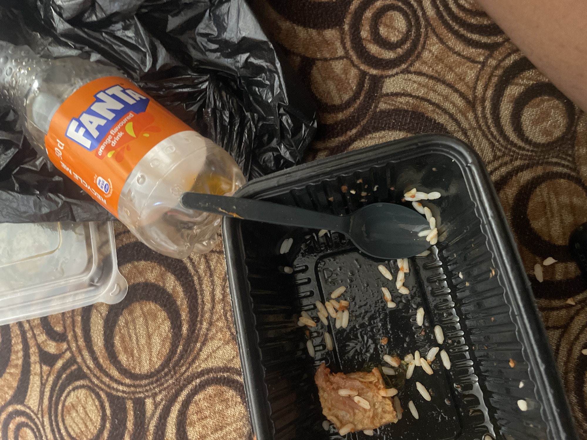 A close-up, overhead shot depicts the aftermath of a meal on a brown, circular patterned carpet. In the foreground, a black plastic food container holds a small portion of white rice and a piece of what appears to be fried chicken. A black plastic spoon rests inside the container. To the right of the container, a partially consumed bottle of orange Fanta, labeled "50 cl" and "orange flavoured drink," lies on its side. Behind the bottle, a crumpled black plastic bag is visible. The overall scene suggests a casual, possibly outdoor setting, given the carpet's presence. The image does not contain information about Lokoja, Nigeria.