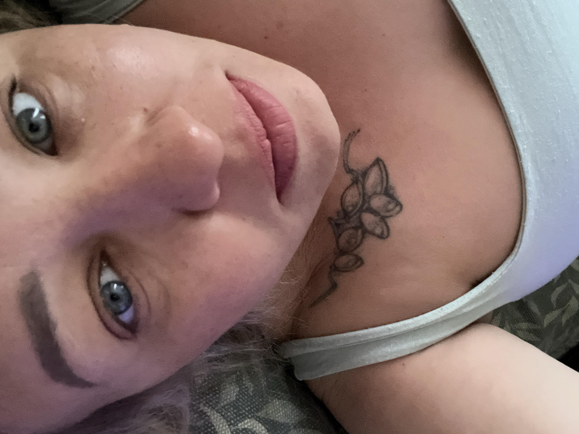 A close-up, slightly angled view depicts a fair-skinned person with gray eyes and light brown hair. They are wearing a light-colored tank top. A dark, stylized floral tattoo is visible on their upper chest/shoulder area. The background is blurred, showing a patterned surface, possibly fabric. The location of the photo, Chaikovsky, Russia, is not evident from the image itself.