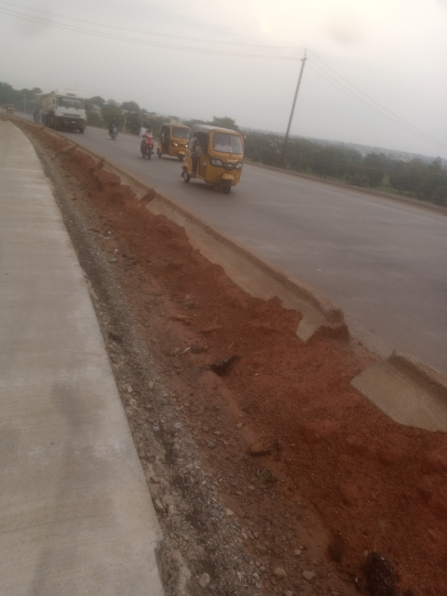 In Goni Gara, Nigeria, this image captures a section of a road under construction. The foreground features a newly laid concrete sidewalk adjacent to a trench filled with reddish-brown soil and gravel. Beyond the trench, the asphalt road is visible, with several vehicles present. A white truck is seen in the distance, moving away from the camera. Closer to the foreground, two yellow auto-rickshaws are on the road, one with a person visible inside, and another with a person riding on the back. Several motorcycles are also present on the road, some with riders. The background shows a line of trees and a hazy sky, suggesting an overcast day. A utility pole with wires stands near the road, indicating the presence of infrastructure. The overall scene depicts a typical day on a developing road network.