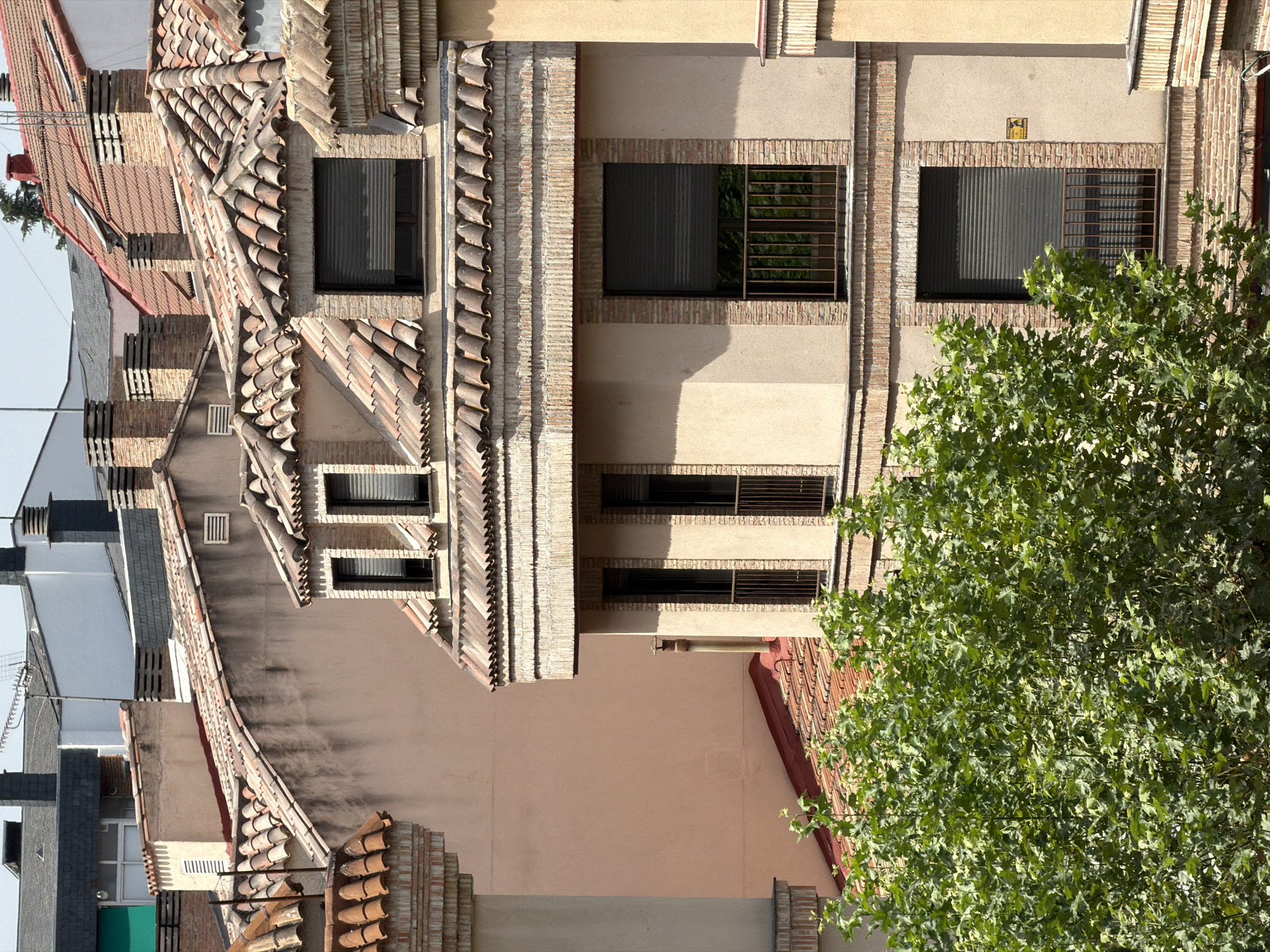 A close-up, eye-level, vertical shot captures the upper floors of several residential buildings in Las Rozas de Madrid, Spain. The buildings feature terracotta tiled roofs with exposed brick chimneys. The facades are predominantly a pale peach color, accented with brick detailing around windows and cornices. Several windows are visible, some with dark shutters partially or fully closed, and some with narrow, dark metal railings on small balconies. Lush green foliage from a large tree dominates the lower left quadrant of the image, partially obscuring the lower levels of the buildings. The sky is a pale blue and appears clear. The architectural style suggests a European urban setting.
