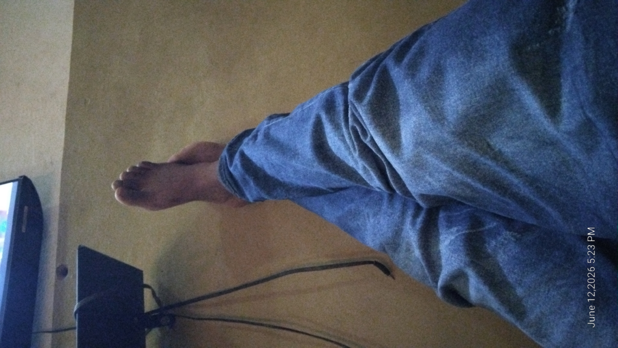 The image is a point-of-view shot of a person's legs and feet, dressed in blue jeans, with their legs crossed. A television screen is visible in the upper left corner, and a black stand with cables is below it. The setting appears to be indoors, likely a living room, on a carpeted floor. A timestamp "June 12, 2026 5:23 PM" is present in the lower left corner. The specific location in Howayi, Nigeria is not determinable from the visual information provided.