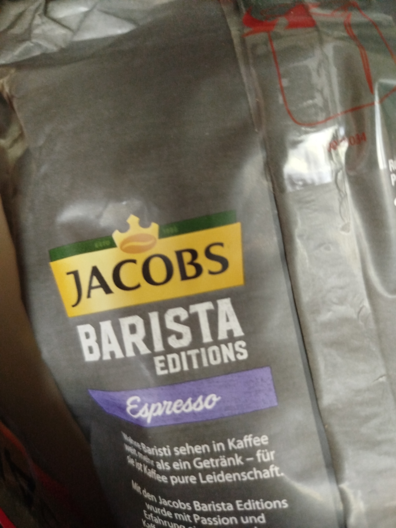 The image shows a close-up of a bag of Jacobs Barista Editions Espresso coffee. The bag is predominantly gray with yellow and green branding. The Jacobs logo, featuring a crown and coffee bean, is prominent. Below that, "BARISTA EDITIONS" is written in large white letters, followed by "Espresso" in stylized purple text. Some smaller text is also visible at the bottom of the bag, likely describing the product. The bag appears to be made of a flexible, possibly foil-lined material. The lighting suggests an indoor setting, and the angle of the shot is from above and to the side. There are no people or specific environmental details identifiable that would indicate a location in Крывасельскі сельскі Савет, Belarus or any other specific geographic area.