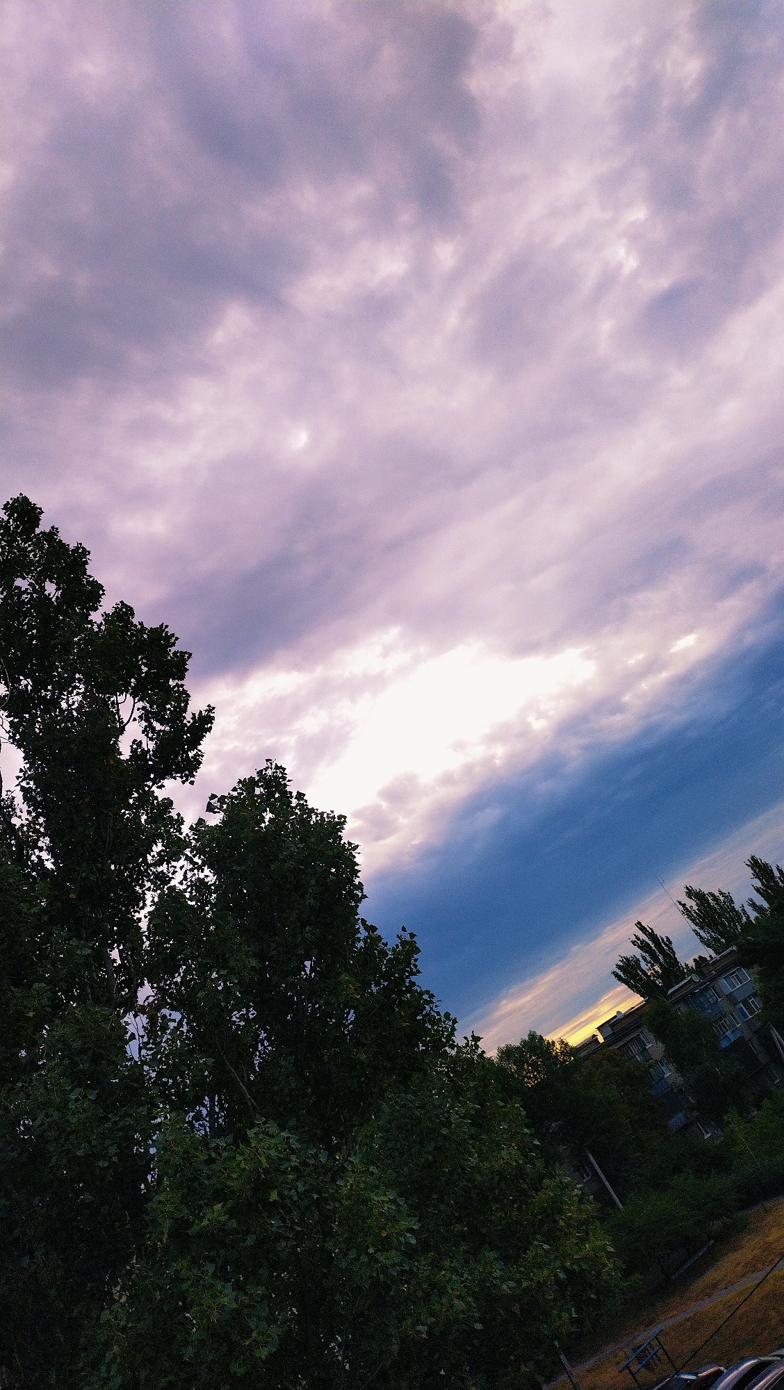 The image depicts a view of a residential area in Horishni Plavni, Ukraine, likely during the late afternoon or early evening, given the lighting conditions. The sky is filled with dramatic clouds, displaying a gradient of colors from soft lavender and pink near the top, transitioning to darker blues and grays as it moves towards the horizon. A bright, hazy patch of light suggests the setting sun is either hidden or just beyond the visible frame.
In the foreground, lush green trees dominate the left and lower portions of the image, their dense foliage obscuring much of the ground below. These trees appear to be deciduous and are in full leaf, indicating a warmer season.
In the middle ground and background, a multi-story apartment building is visible, characterized by its blue and gray facade. Windows are sparsely visible, and the building's architecture suggests a typical Soviet-era design. Surrounding the building are more trees and what appears to be a patch of yellowish grass or dry vegetation, possibly a courtyard or a small park area. The presence of a utility pole or antenna is also noted.
The overall perspective is from a slightly elevated viewpoint, looking out over the trees and towards the residential buildings. The composition is diagonally bisected by the sky and the tree line, creating a dynamic visual flow. No people are visible in the frame, and no specific actions or interactions are depicted. The scene conveys a sense of urban nature juxtaposed with everyday residential life, under a striking sky.