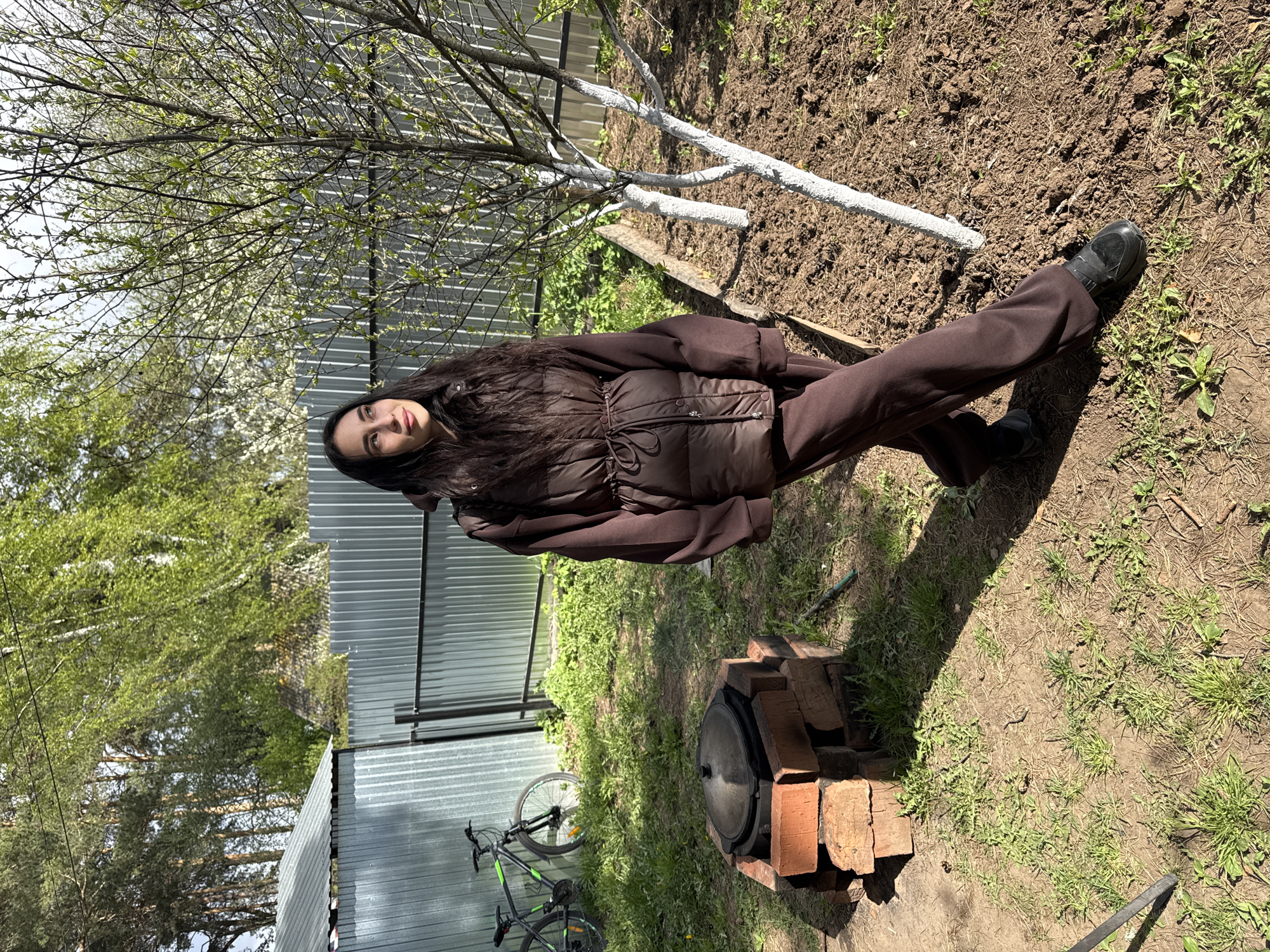 A young woman stands in a garden in Chaikovsky, Russia. She wears a brown jacket and matching trousers with black shoes. Behind her, a freshly painted white tree trunk rests on a patch of bare earth. In the background, there is a grey metal fence and a bicycle is parked near a structure made from bricks with a black pot on top. Green trees surround the scene, suggesting a rural or suburban environment. The weather appears to be sunny, casting distinct shadows on the ground.