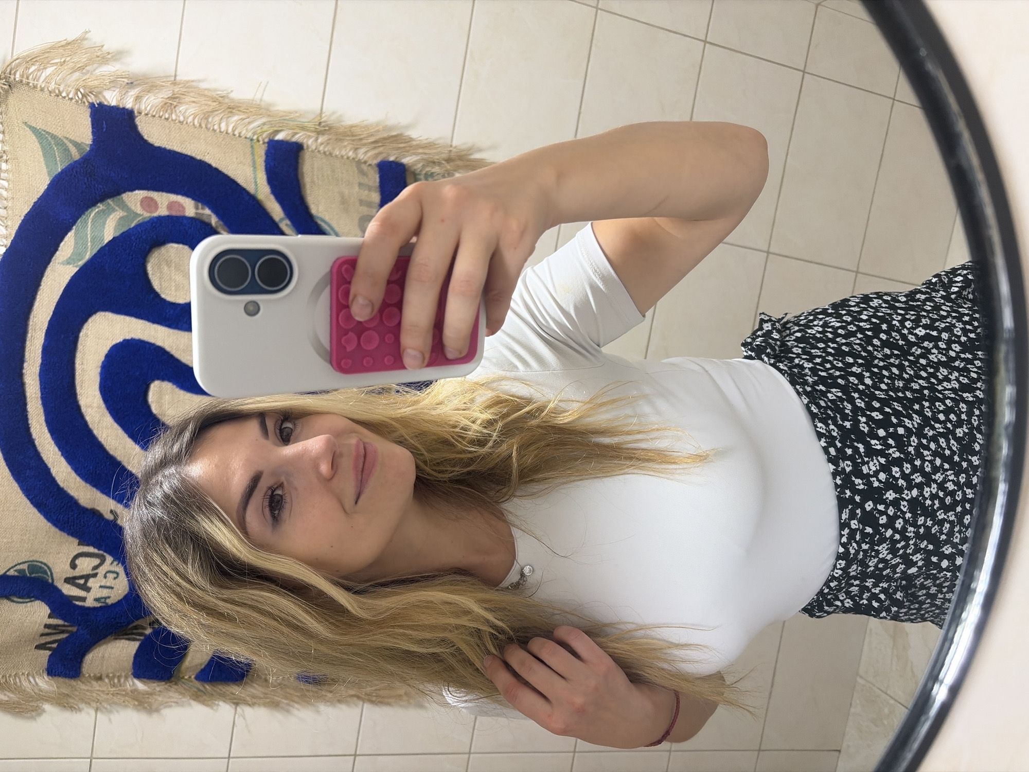 A woman is taking a mirror selfie in what appears to be a bathroom. She has blonde, wavy hair and is wearing a white t-shirt and a black skirt with a floral pattern. She is holding a white smartphone with a pink pop socket in her right hand to take the photo. In the background, a tiled wall is visible, and above it, a tapestry with blue and beige designs hangs. The overall setting suggests an indoor, personal space, potentially a home or hotel bathroom in Budva, Montenegro.