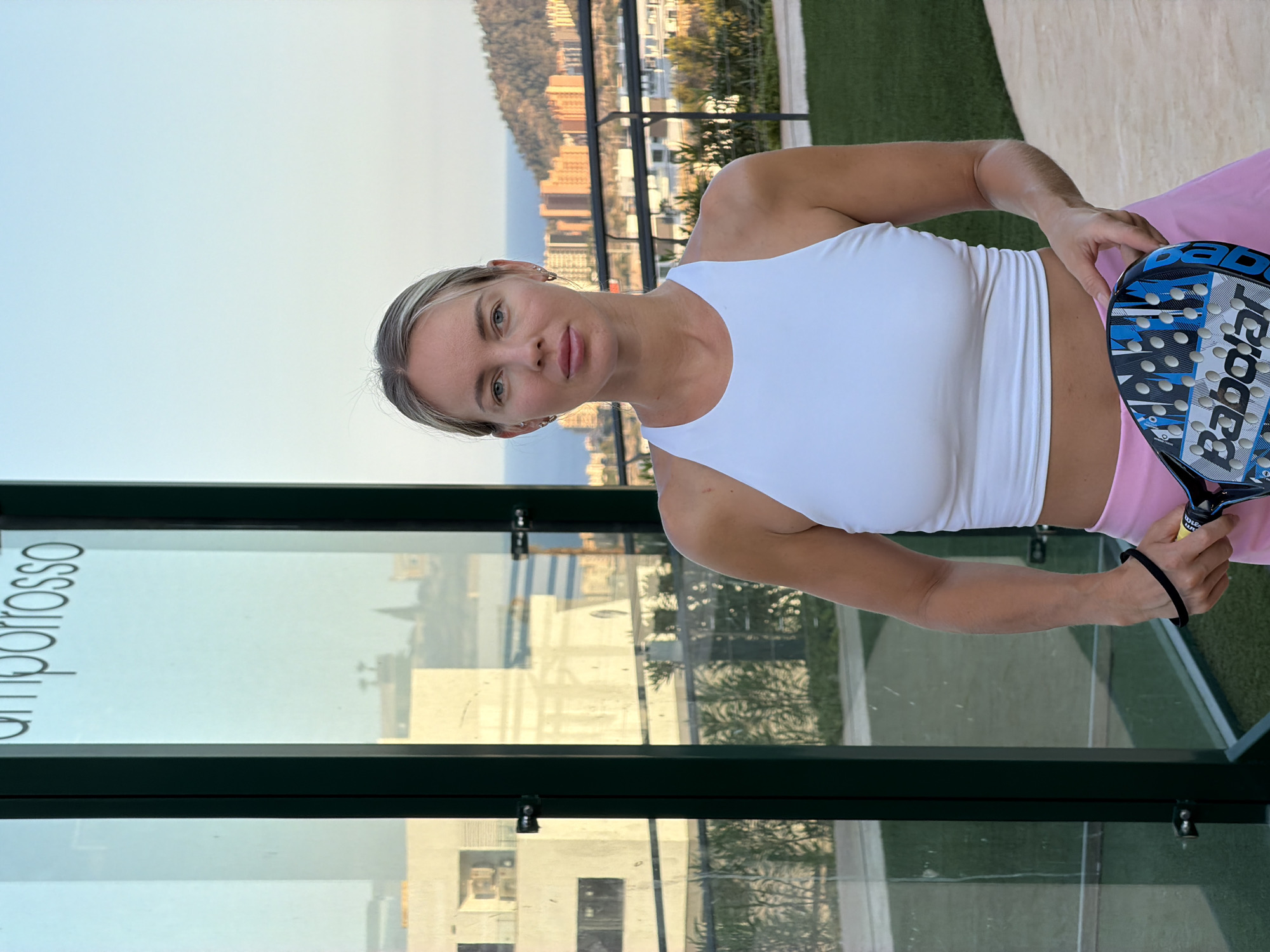 A blonde woman wearing a white sports top and pink skirt stands in front of a glass structure with the word "emporiosso" visible on it. She is holding a blue and black paddle tennis racket. In the background, there is a cityscape with buildings, a mountain, and the sea under a clear sky. The location is identified as Finestrat, Spain.