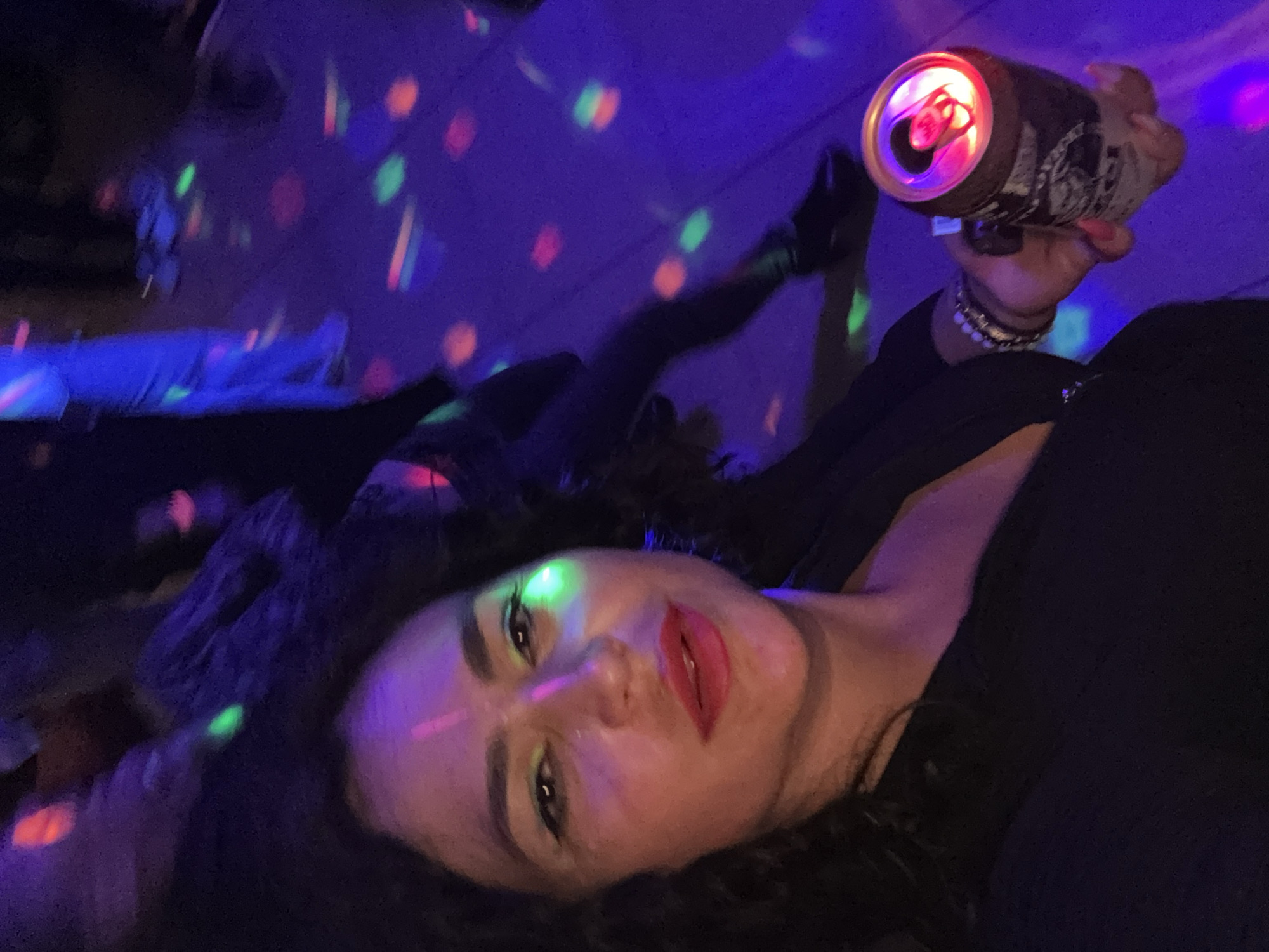 A woman lies on the floor in a darkened space with colored disco lights projected onto the walls and floor. She is holding an open can of beer in her right hand. The room appears to be indoors, based on the artificial lighting. Other figures are visible, though less distinct. The specific location within Chaikovsky, Russia is unclear without further context.