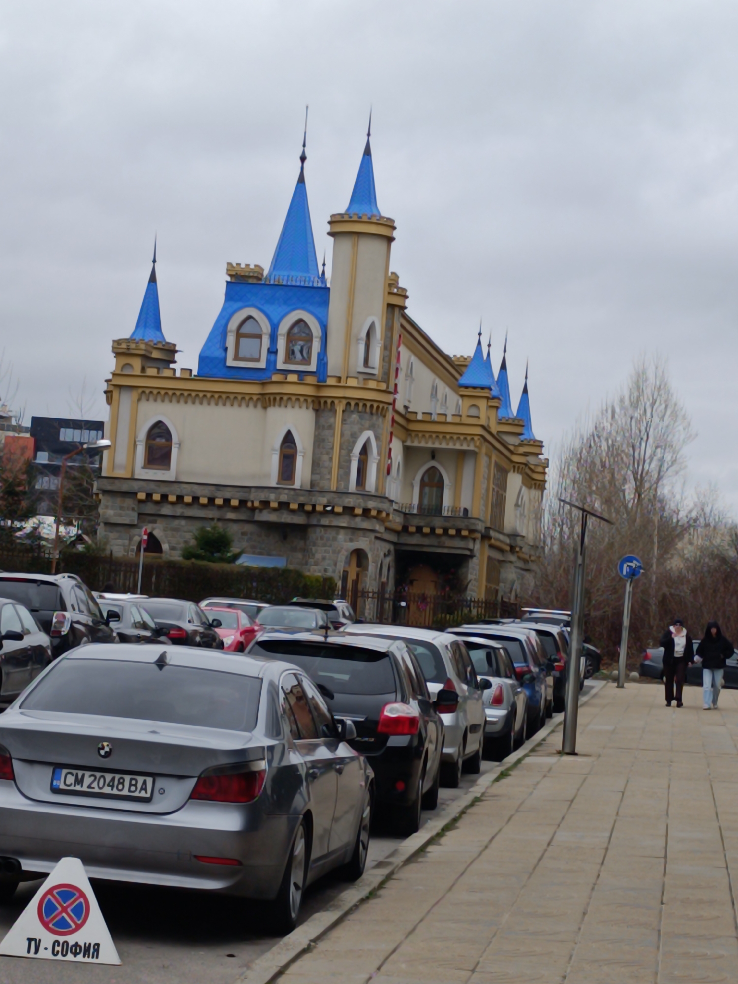 The image shows a row of parked cars along a sidewalk in front of a building resembling a castle. The building has a light beige facade with stone accents, blue turrets, and ornamental trim. A "no parking" sign stands on the sidewalk near a silver car with the license plate "CM 2048 BA." Two people are walking along the sidewalk away from the camera. The location appears to be in Sofia, Bulgaria, based on the language on the "no parking" sign. The sky is overcast.
