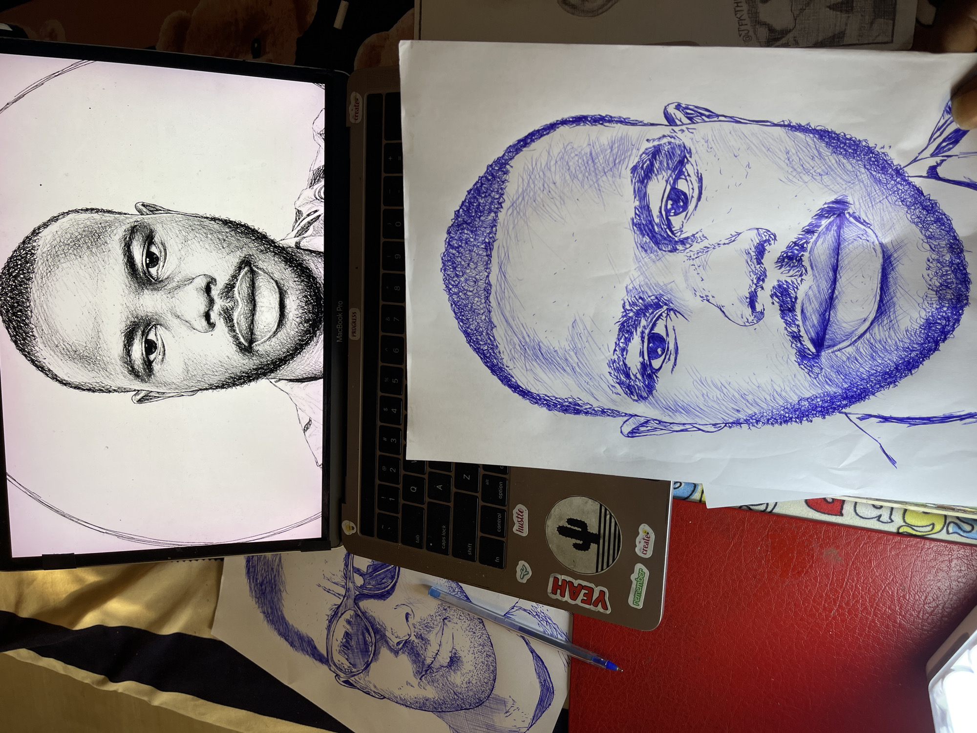 The image depicts a desk setting with artwork and a laptop, likely in an indoor environment. The primary focus is on two blue ink drawings of a man's face. One drawing is displayed on the screen of an open MacBook Pro laptop, showing a close-up portrait with detailed shading. Another, more complete drawing of the same man's face is laid out on a white sheet of paper in the foreground, also rendered in blue ink with extensive cross-hatching for shading. Next to the laptop, another drawing of a man wearing sunglasses is partially visible. A blue ballpoint pen rests on top of this drawing and the laptop's keyboard. The laptop has several stickers on it, including one with a cactus illustration and text like "YEAH," "hustle," "remember," and "create." The desk surface appears to be red and textured. The background includes a striped fabric and what looks like a stuffed teddy bear. There is no identifiable information or context within the image that would allow for the specific determination of its location in the city of Jos, Nigeria.
