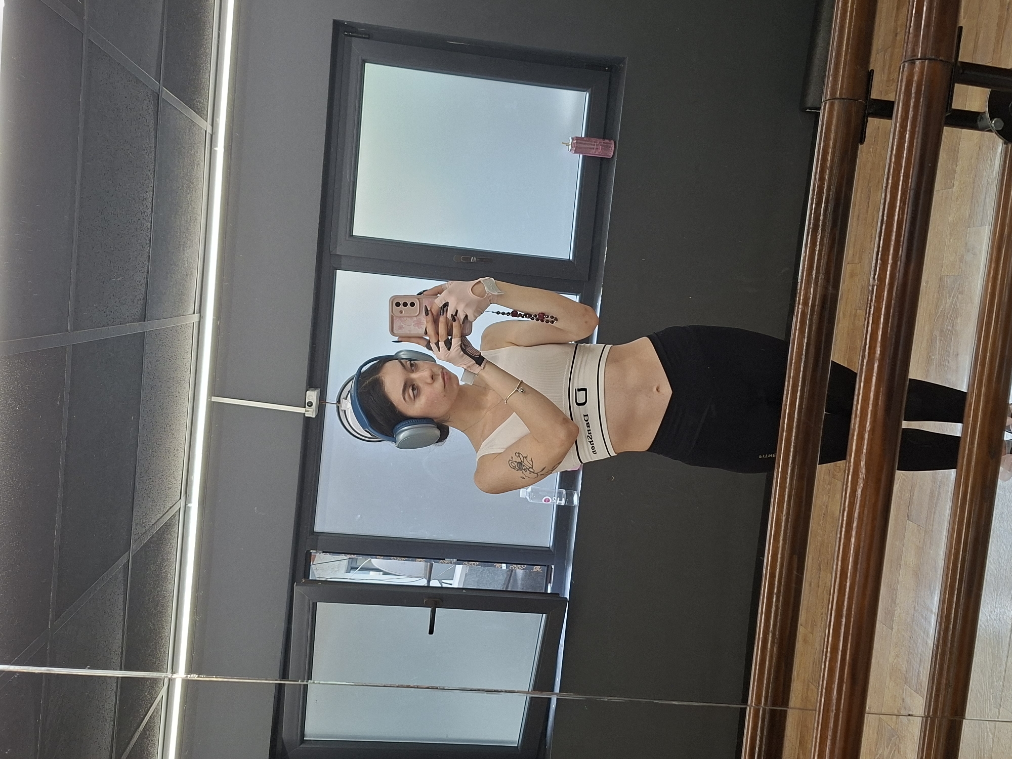 An image, captured in Sofia, Bulgaria, shows a young woman taking a mirror selfie within what appears to be a modern fitness or dance studio. The woman is positioned centrally, holding a light pink smartphone with a decorative case to photograph her reflection. She is wearing light blue over-ear headphones, a white crop top or sports bra with a prominent band, and black leggings. Her hands are adorned with light pink fingerless gloves, and a thin bracelet is visible on her left wrist. A small, abstract tattoo is visible on her right forearm. She has dark hair and a calm expression, looking towards her phone screen.
Behind the woman, large windows allow natural light into the space, and a pink water bottle, clearly marked "H2O," rests on the window ledge. The studio's interior features dark grey walls, some of which have textured paneling, and a ceiling equipped with bright, linear LED strip lighting. To the right, a set of polished wooden poles, indicative of a ballet barre or similar exercise equipment, is reflected in the mirror. The floor, visible as a reflection beneath the woman, is made of light-colored wooden parquet. The white band on her crop top displays mirrored black text that reads "DЯƎMИИD" (DREAMING when unmirrored), and faint mirrored text, possibly "ELLITE," is visible on her leggings. The scene suggests a daytime setting within an active environment.