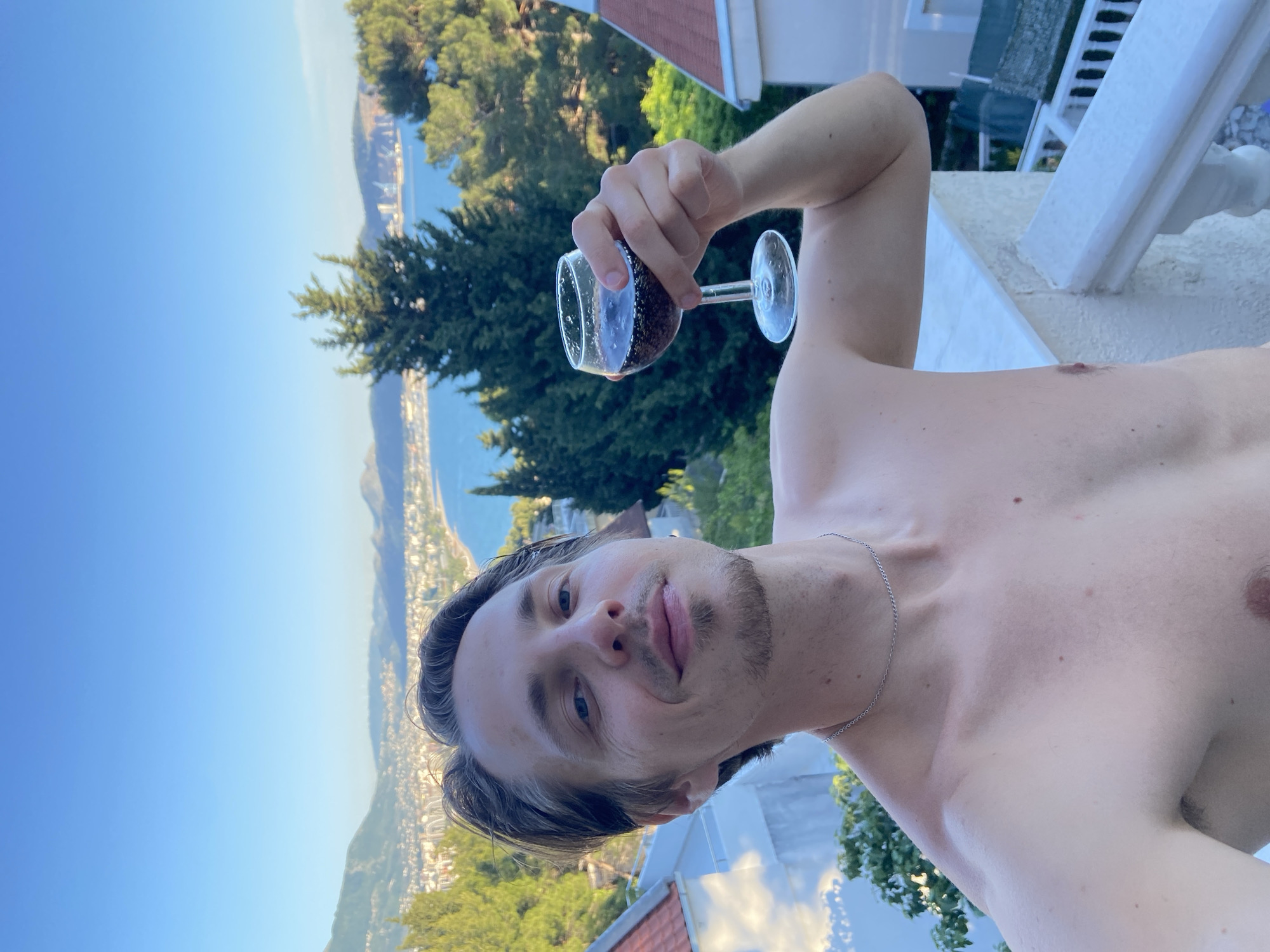 A shirtless Caucasian male with short brown hair and stubble holds a wine glass containing a dark liquid. He is standing on a white concrete balcony or terrace. In the background is the city of Šušanj, Montenegro, situated along the coastline, with clear blue sky above. There are green trees and rooftops visible in the surrounding area. The man is facing the camera with a slight smile. A silver necklace is visible around his neck.