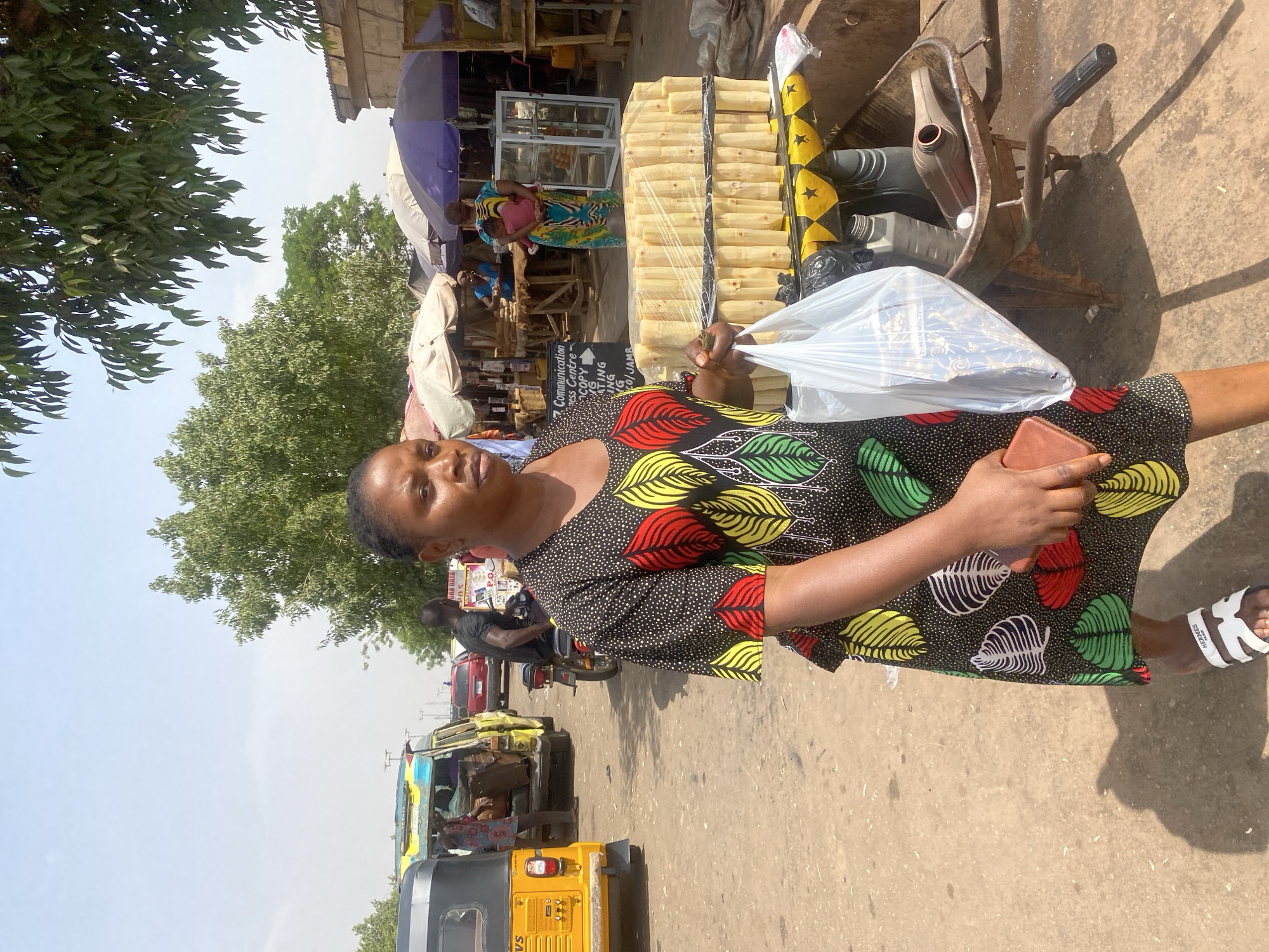 The scene depicts a bustling outdoor market area in Sabon Tasha, Nigeria, under clear daylight.
In the foreground, a woman with dark skin and braided hair stands slightly off-center to the left, facing generally rightward but looking slightly left. She wears a black, short-sleeved dress with a pattern of red, yellow, green, blue, white, and purple leaves with white dots. Her left hand holds a white plastic shopping bag containing items, and her right hand holds a brown mobile phone. She wears white slides with "HERMES" branding. Her shadow is cast on the dirt ground to her right.
Immediately to her right, a modified, rusty wheelbarrow serves as a mobile display unit. It is covered with a yellow and black striped fabric featuring stars, resembling the flag of Ghana. Stacked on top of this wheelbarrow are numerous light-colored, cylindrical items, likely sugarcane pieces, wrapped in clear plastic and secured with black tape. Beneath this stack, inside the wheelbarrow's basin, are various plastic bottles, some containing liquids. A small black-and-white sign on the wheelbarrow advertises "Communication Centre" services including "COPY," "TYPING," "TING," "ING," and "JAMB," with an arrow pointing right.
In the midground, an unpaved road occupies the left and central areas. A yellow motorized tricycle (TVS brand) is visible on the far left, facing away from the viewer. To its right, a yellow van is partially visible, with a person in a colorful shirt near its rear. Further right, a man in a dark shirt is on a red motorcycle, facing away. Along the right side of the road, a series of market stalls are set up, covered by assorted parasols in white, brown, and purple hues. Another woman, wearing a blue and yellow patterned dress, holds an infant clad in pink, facing the stalls. Partially visible signs read "DATA," "50 S," and "P.C.O."
The background reveals more green trees and distant, low-rise buildings. The sky is a light blue with some haze. Shadows are distinct, indicating bright sunlight. Other vehicles, including a red car, are visible further down the road.