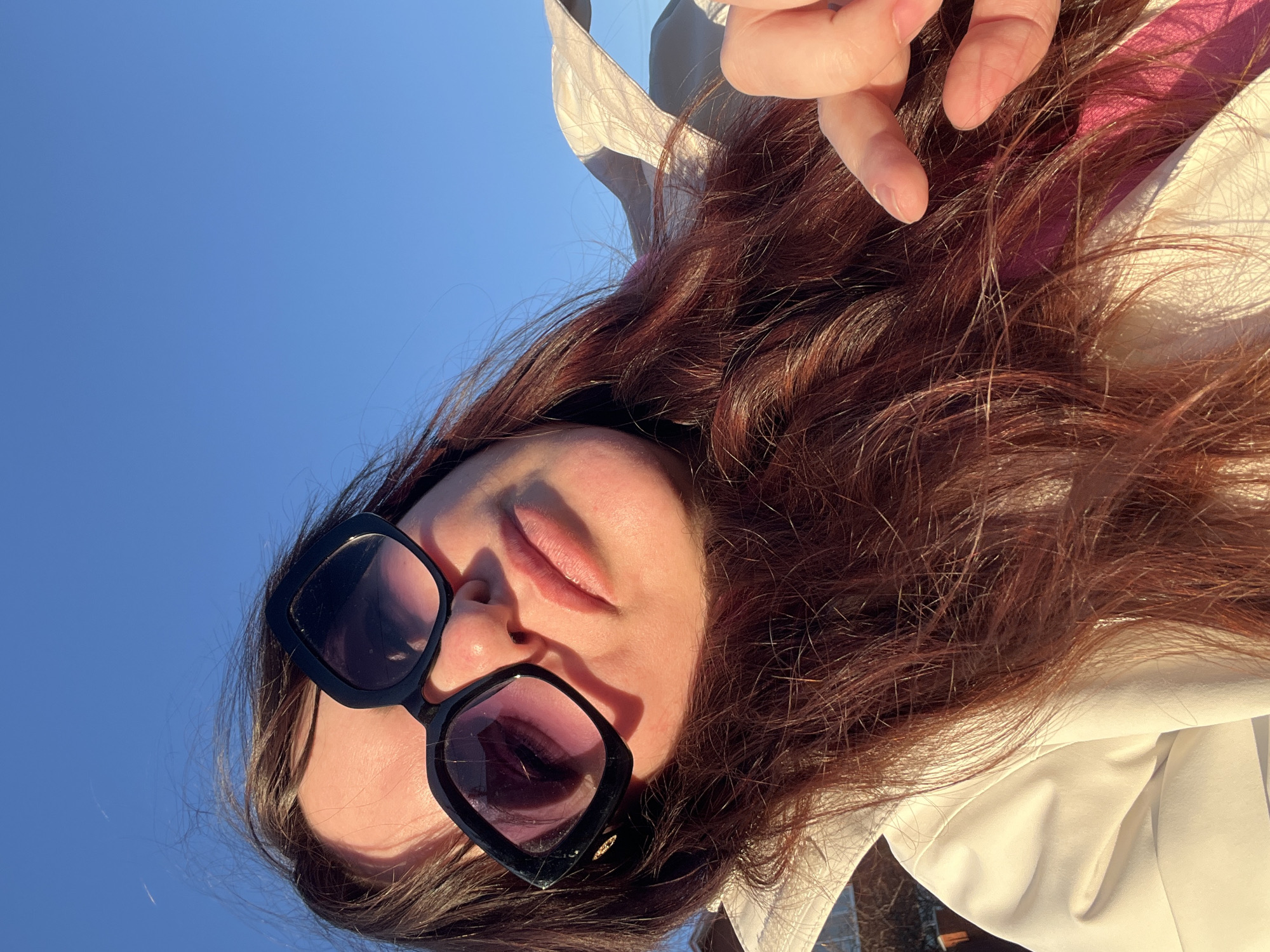 A low-angle selfie of a person with long, reddish-brown hair wearing black sunglasses against a clear blue sky. The person's face is partially visible, showing closed eyes, nose, and lips. A hand with fair skin and trimmed fingernails is near the top of the frame. The person is wearing a white top or jacket. The exact location in the city of Noviy, Russia, cannot be determined from the image.