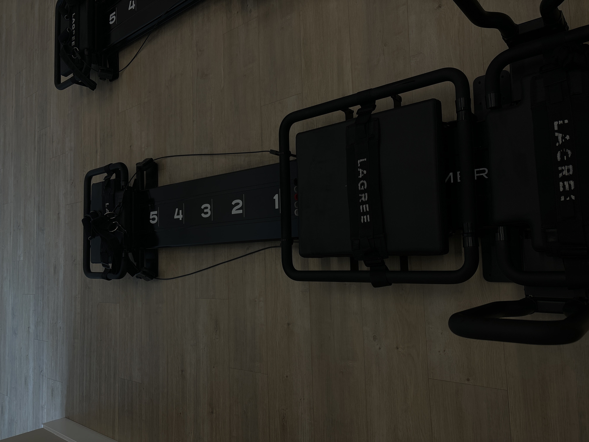 The image shows two Lagree fitness machines on a wood-look floor. The machines are black and feature numbered indicators from 1 to 5. The words "Lagree" and "MER" are visible on the machines. The location appears to be indoors, potentially in a fitness studio.