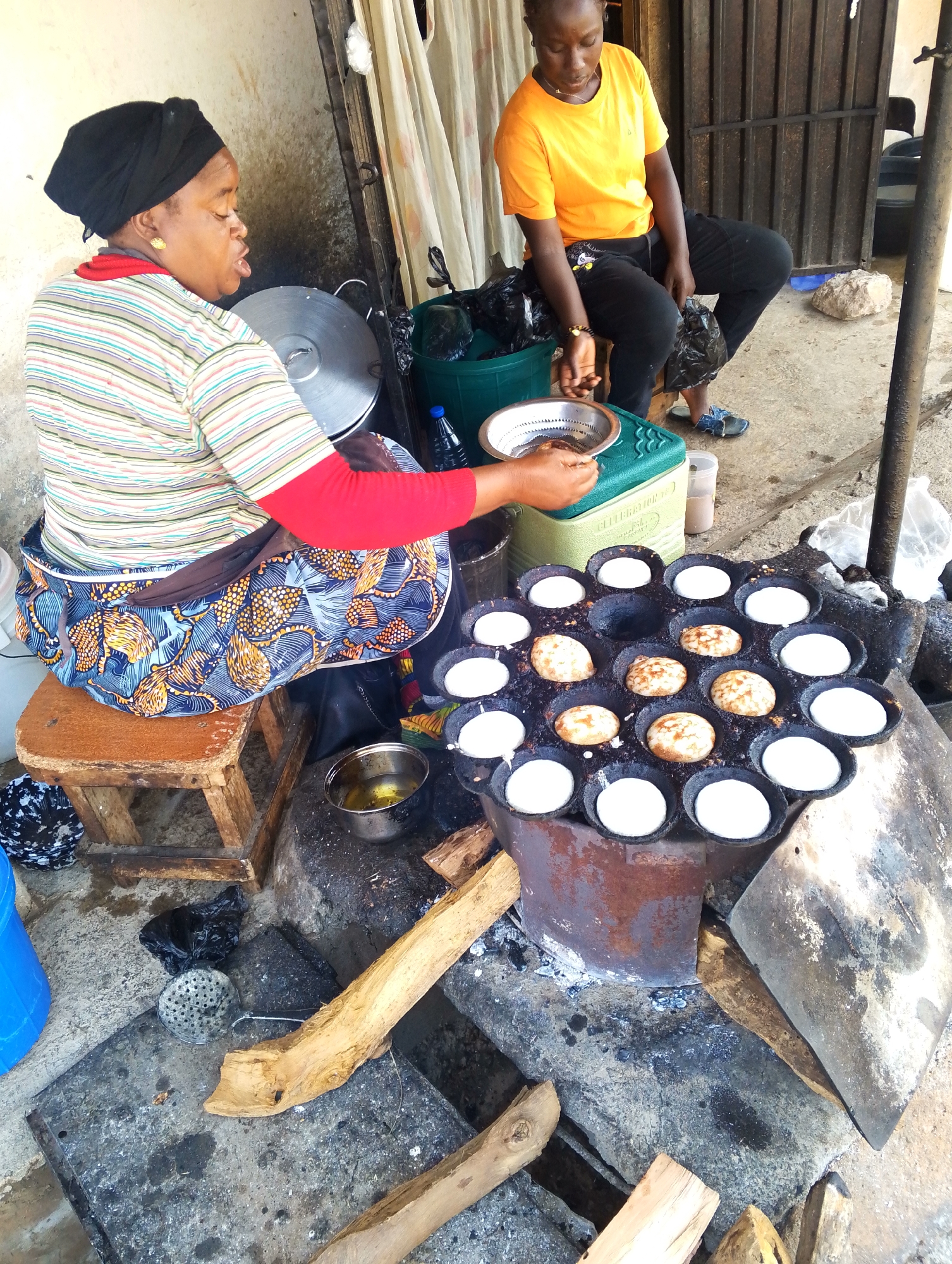 This is a medium shot, taken outdoors, of two women preparing food over a fire in Jos, Nigeria. In the foreground, a woman wearing a black headwrap, a striped shirt, and a blue and yellow patterned apron is seated on a wooden stool, tending to a specialized pan with multiple round indentations where small cakes are being cooked over an open fire. The cakes are in various stages of cooking; some are white, while others have browned edges. Next to the cooking pan is a metal bowl containing oil, and several pieces of firewood. In the background, a second woman wearing an orange t-shirt and black pants is seated and holding a plastic bag. Behind them, a doorway with a metal grate is visible, as are plastic containers and various pieces of debris. The overall environment appears to be an informal outdoor cooking or market stall area.
