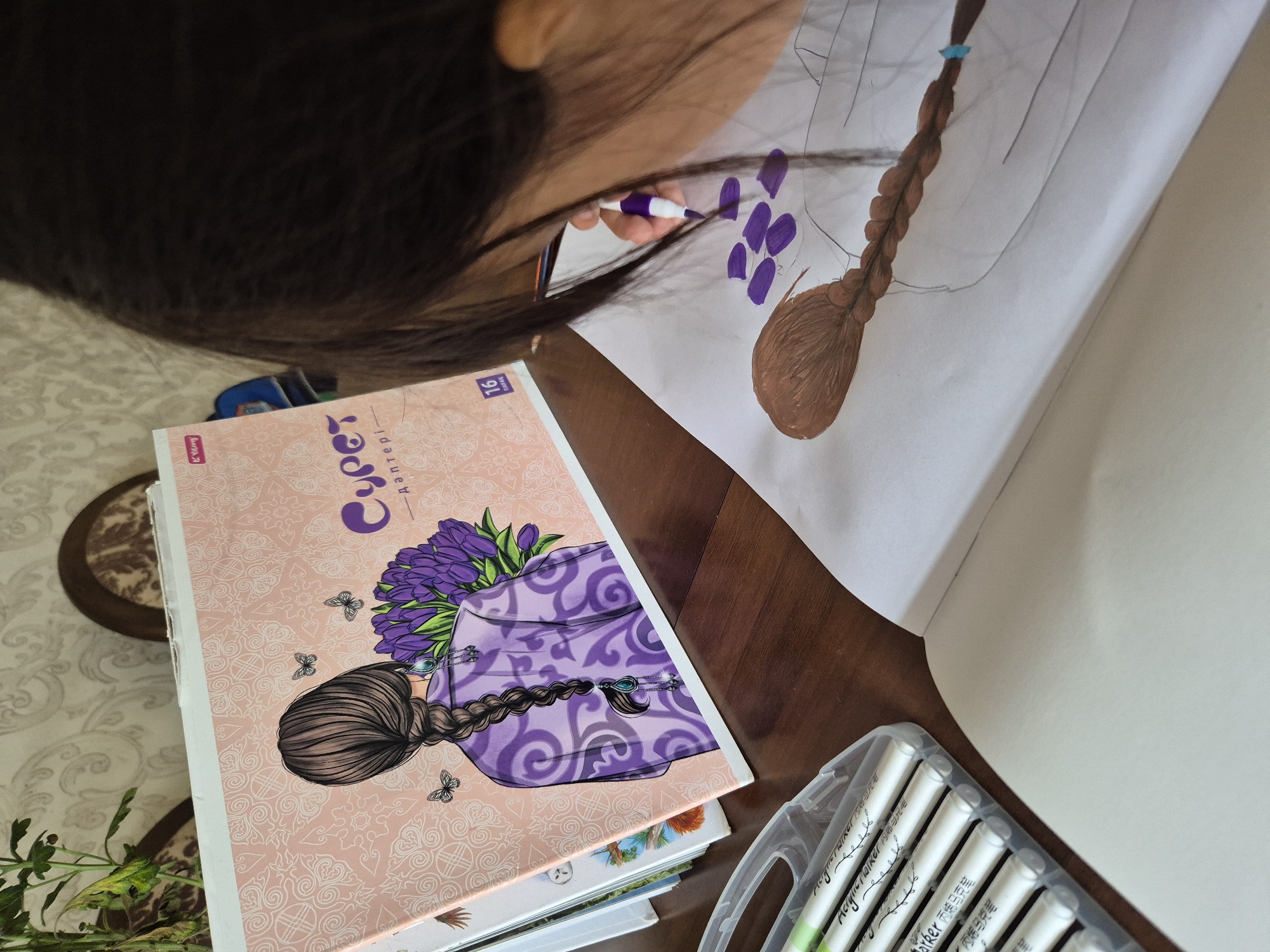 A young girl with dark hair, seen from behind, is drawing on a piece of paper with a purple marker. She is sitting at a wooden table. In front of her is an open art book with an illustration of a woman with a braid and flowers. To her left, a set of white markers in a clear plastic case is on the table. The background shows a patterned wallpaper and a chair. The scene is indoors, likely in a home, and there is no specific indication of the city of Kokshetau, Kazakhstan.