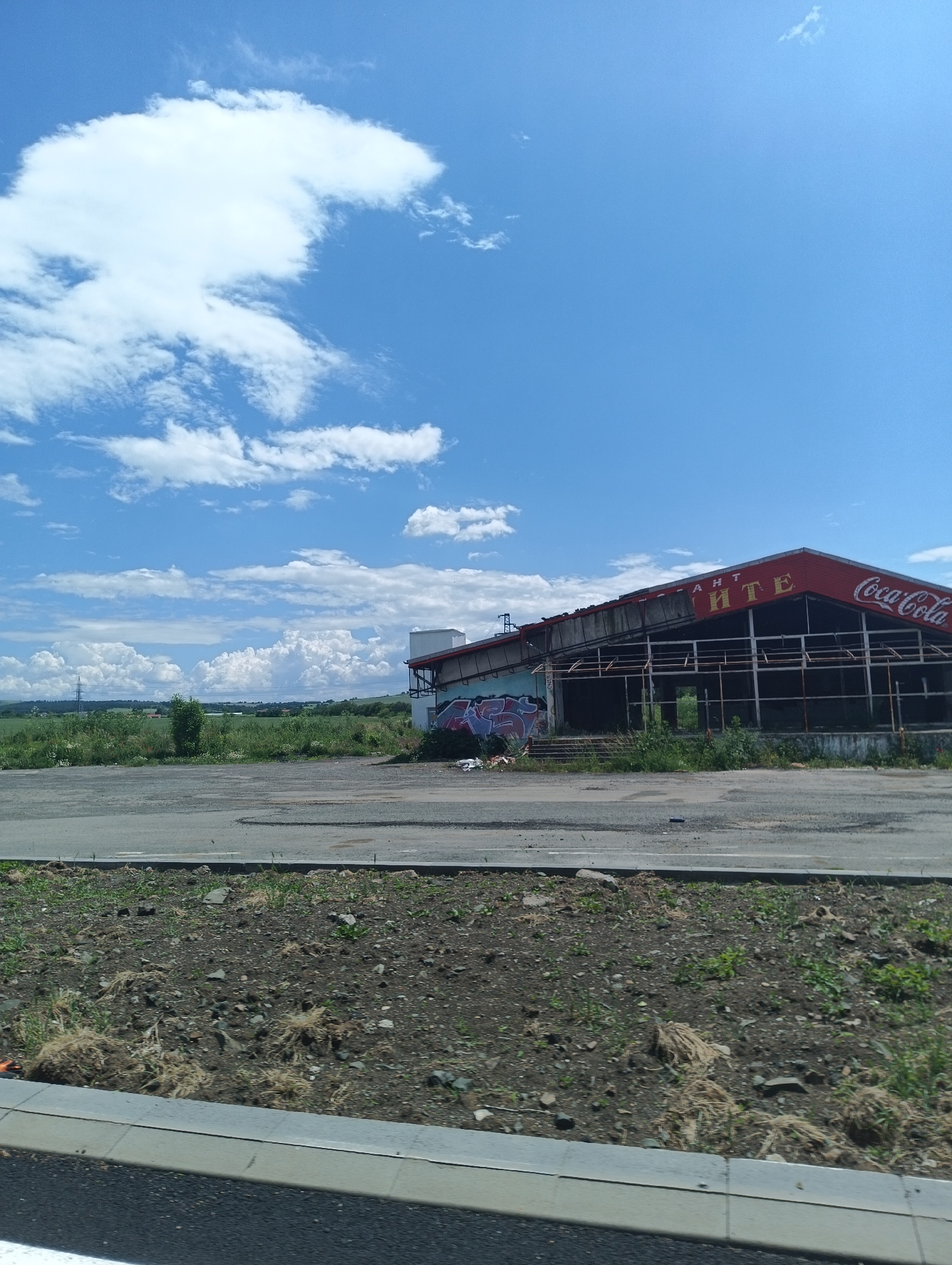 The image shows a dilapidated, abandoned building located near Sokolovo, Bulgaria. The building's red roof displays faded signage with the Coca-Cola logo and the word "PHTE" partially visible. The structure is mostly bare framing with evidence of graffiti on its exterior. The building is positioned next to a paved area that appears cracked and worn. In the background, there is a field of green vegetation and cloudy skies. The foreground consists of a grassy area with a curb in front.