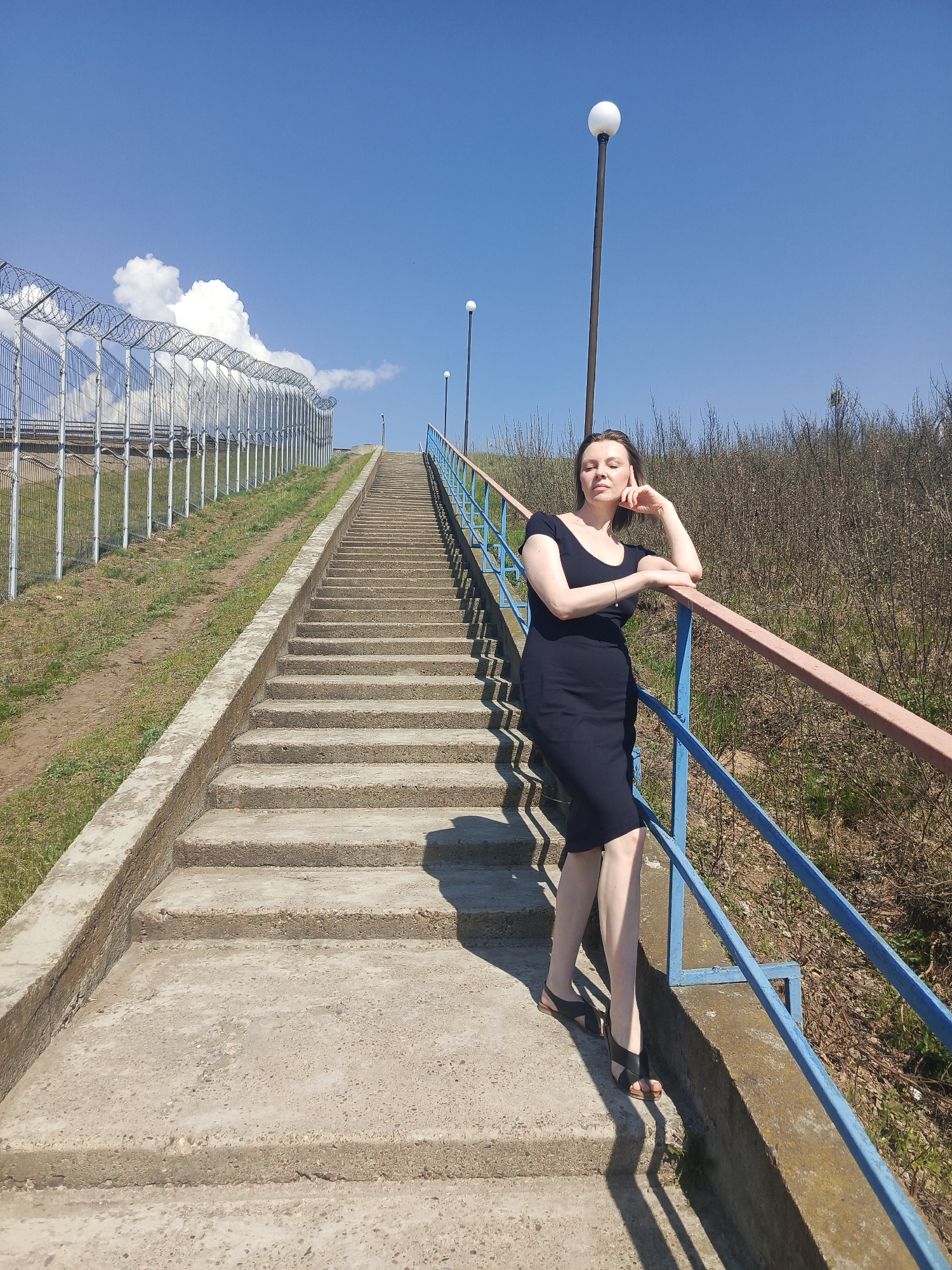 A full shot captures a woman standing on a long outdoor staircase with blue handrails. The staircase ascends a slope, with a chain-link fence topped with barbed wire running parallel on the left. On the right, the slope transitions into scrubby vegetation. The woman is dressed in a knee-length black dress and black sandals, leaning against the handrail with her right hand resting on her face. Behind her, evenly spaced light poles rise along the staircase. The sky is clear blue with some fluffy white clouds near the top of the fence. The scene is bathed in sunlight, casting distinct shadows. The location appears to be a public walkway in an urban environment, specifically Unknown, Unknown.