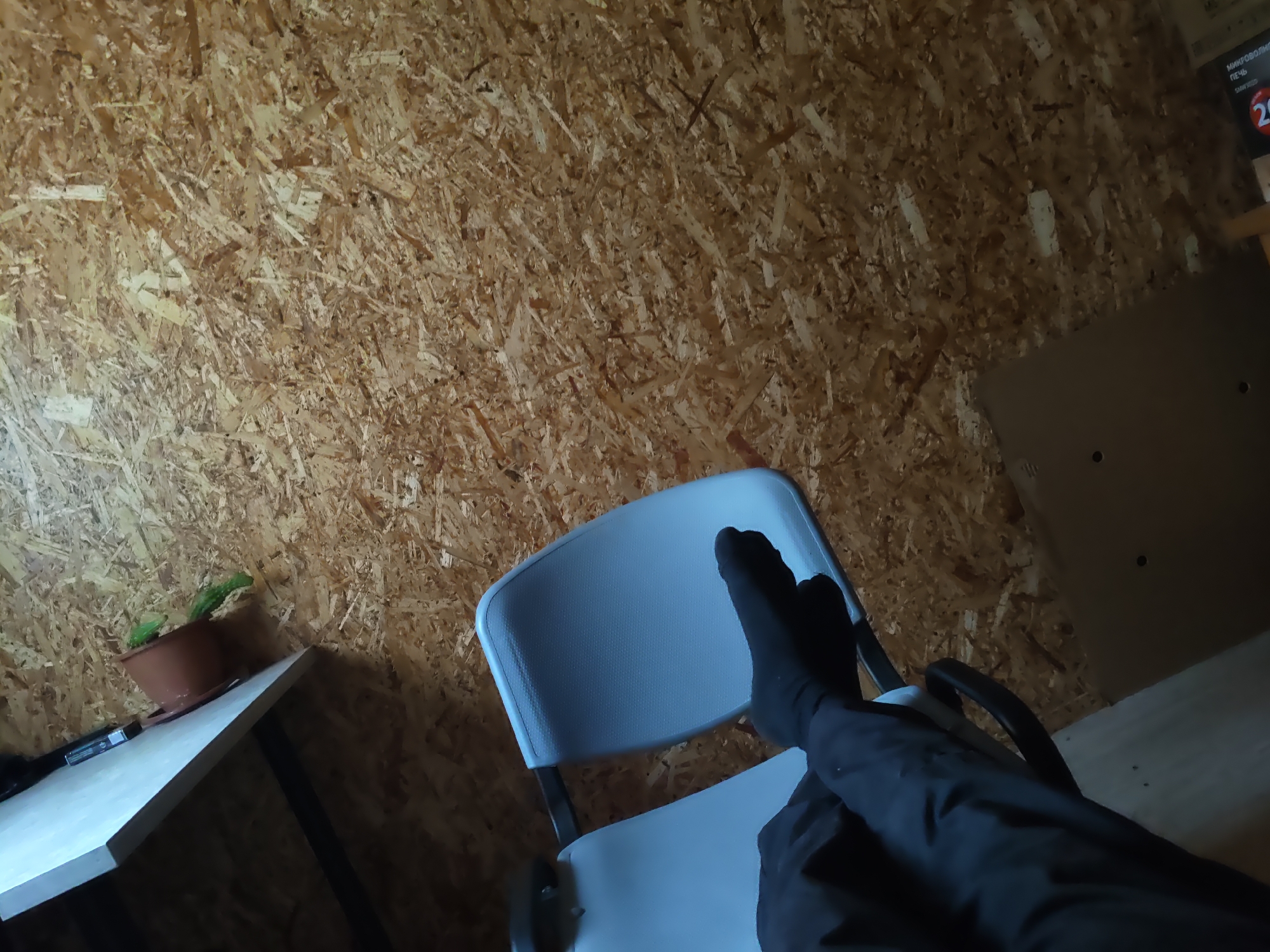 The image depicts an indoor scene with a person's legs, wearing black socks, resting on a white plastic chair. The chair is positioned in front of a wall constructed from oriented strand board (OSB), which has a rough, textured surface. To the left, a light-colored table is visible, and on it sits a brown pot containing a green plant, possibly a cactus. The room appears to be dimly lit, with the OSB wall occupying the majority of the background. A cardboard box or panel is partially visible on the right side of the frame, and above it, a portion of what appears to be product packaging with Russian text, including the word "ПЕЧЬ" (oven), is discernible. The scene is likely a domestic or workshop setting within the city of Platoshino, Russia.