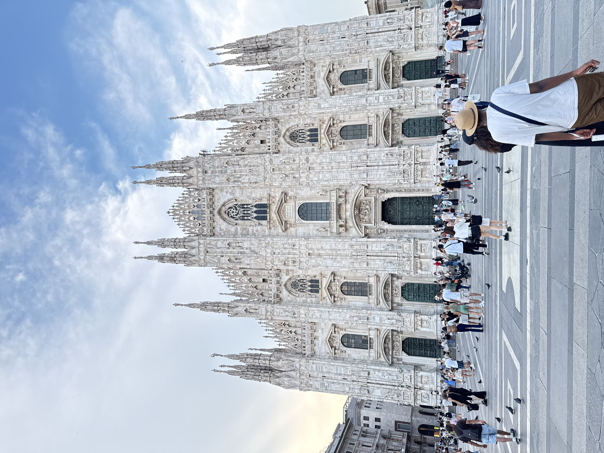 The image depicts the Milan Cathedral, also known as Duomo di Milano, in Milan, Italy. The grand Gothic cathedral, constructed primarily of white marble, dominates the frame with its intricate facade, numerous spires, and arched windows. In the foreground, a paved piazza is filled with a crowd of people. Tourists are seen walking, standing, and observing the cathedral. Some are dressed in casual summer attire, and several are carrying bags or taking pictures. Pigeons are scattered across the plaza. The sky above is a mix of blue with scattered clouds, indicating daytime. To the left of the cathedral, other buildings of the city can be seen. The overall scene captures a busy, public space in front of a major architectural landmark.