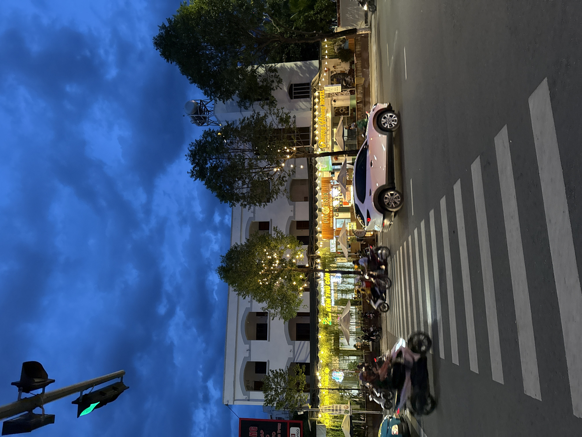 The image depicts a street scene at dusk in Nha Trang, Vietnam. A pedestrian crosswalk dominates the foreground, with several motorcycles in motion crossing it. To the right, a light-colored SUV is also on the road. Behind the crosswalk, a two-story building with arched windows is visible. The building's ground floor features a row of shops or restaurants with awnings and outdoor seating, illuminated by strings of warm white lights. Trees with lights are planted in front of the building. The sky is dark blue with dramatic clouds, suggesting twilight or early evening. A traffic light with a green signal is visible in the upper left corner. The overall impression is of a bustling urban street during a transitional time of day.
