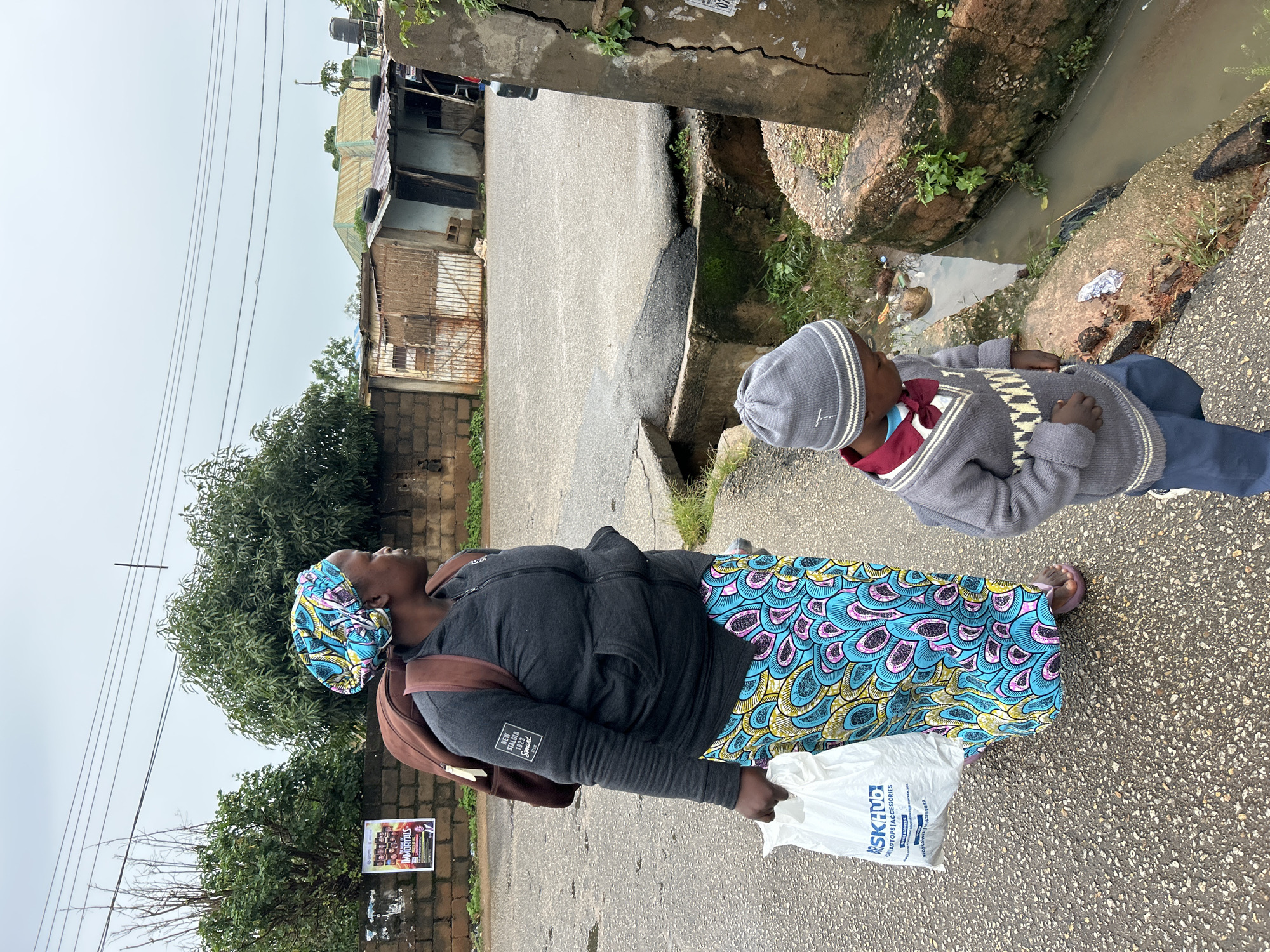 The image shows a medium shot of a woman and a young boy walking on a paved road in Jos, Nigeria. The woman, on the left, is wearing a colorful patterned skirt, a dark gray hooded jacket, and a headscarf. She has a brown backpack on her shoulders and is carrying a white plastic bag in her left hand. The boy, on the right, is wearing a gray knitted hat and sweater, a red shirt with a bow tie, and blue pants. They are walking next to a concrete drainage ditch filled with murky water and debris. In the background, there are brick walls, corrugated metal structures, trees, and a cloudy sky. A poster is visible on a wall to the left, and electrical wires are strung overhead. The scene appears to be in a residential or mixed-use area of the city.
