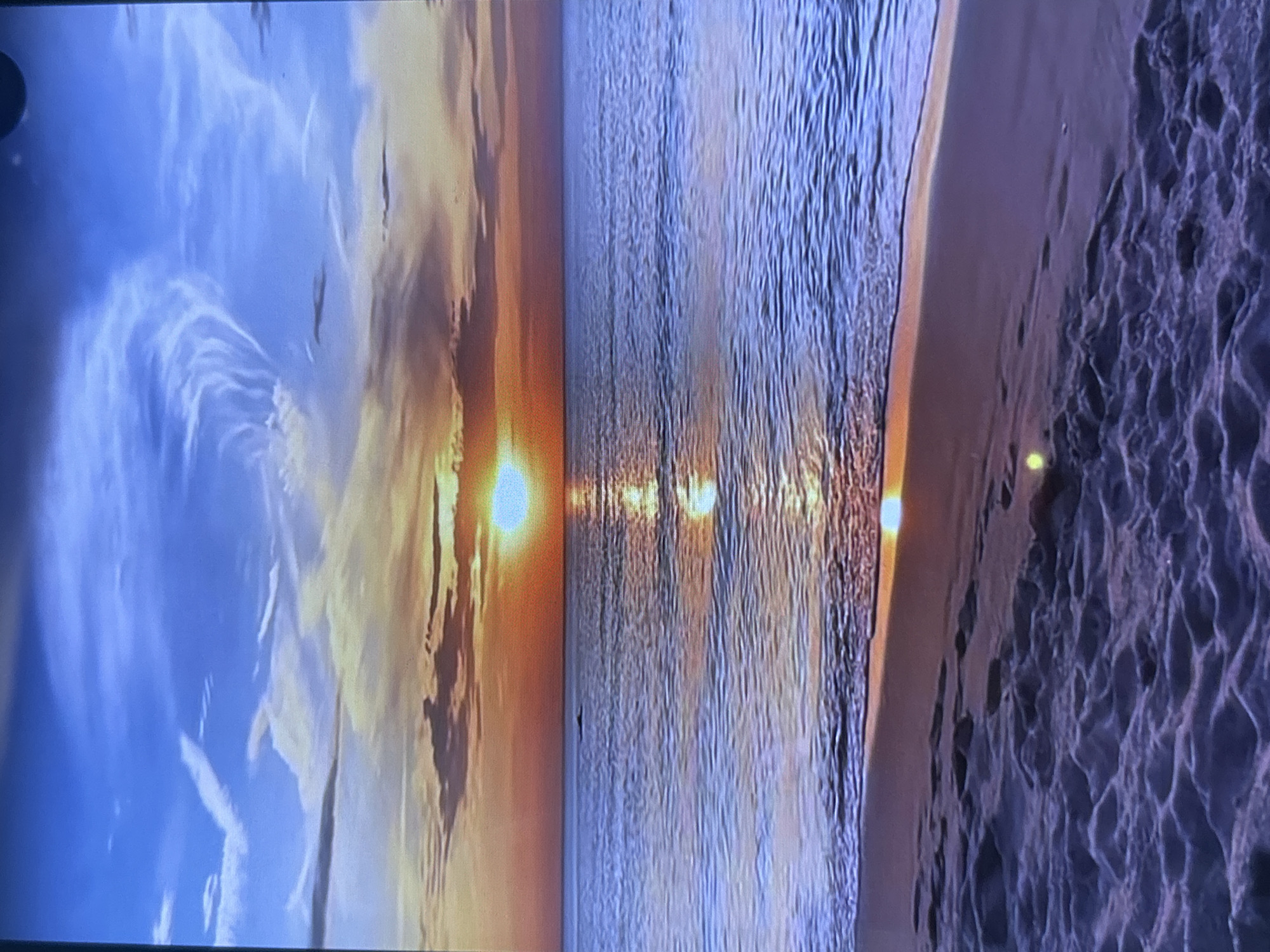 The image displays a serene landscape scene depicting either a sunset or sunrise over a large body of water, likely an ocean or sea, next to a sandy beach.
In the upper portion of the frame, the sky transitions from a soft blue at the top to warm hues of orange and yellow near the horizon. Wispy and scattered clouds are illuminated by the sun's glow, creating a dramatic visual effect. The sun itself is a bright, clear disk, positioned near the horizon line.
Below the sky, the vast expanse of water stretches horizontally across the middle of the frame. The surface of the water is textured with numerous ripples and gentle waves, and it powerfully reflects the sun's light, creating a shimmering path that extends from the sun towards the foreground.
To the right, a sandy beach occupies the lower right portion of the image. The sand exhibits distinct patterns and undulations, suggesting the effects of waves or wind. Small, bright reflections are visible on the sandy surface.
No people or identifiable man-made objects are present in the scene, conveying a sense of untouched natural beauty. A dark, circular artifact, likely a lens flare or part of the camera's lens, is visible in the top left corner of the image. The time of day is clearly either dawn or dusk, characterized by the low sun angle and vibrant sky colors. No visible text is present.
