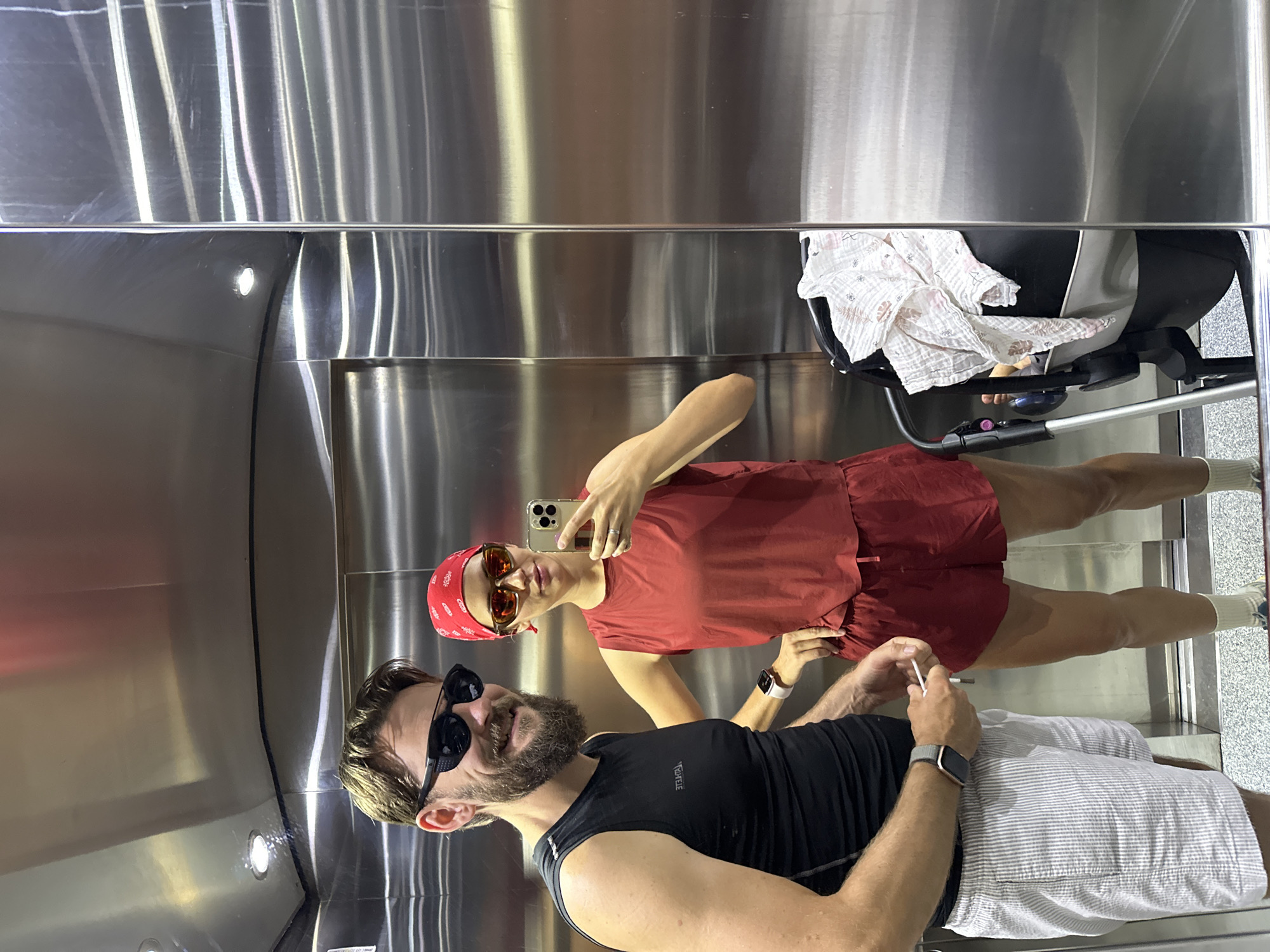 A man and a woman are taking a mirror selfie in an elevator, likely in a hotel in Fethiye, Turkey. The man, wearing a black tank top, sunglasses, and striped shorts, stands on the left. The woman, on the right, wears a red bandana, sunglasses, a red tank top, and red shorts. She is holding a smartphone, taking the photo. A baby stroller is visible to the right of the woman, with a white and pink blanket draped over it. The elevator walls are made of polished stainless steel.
