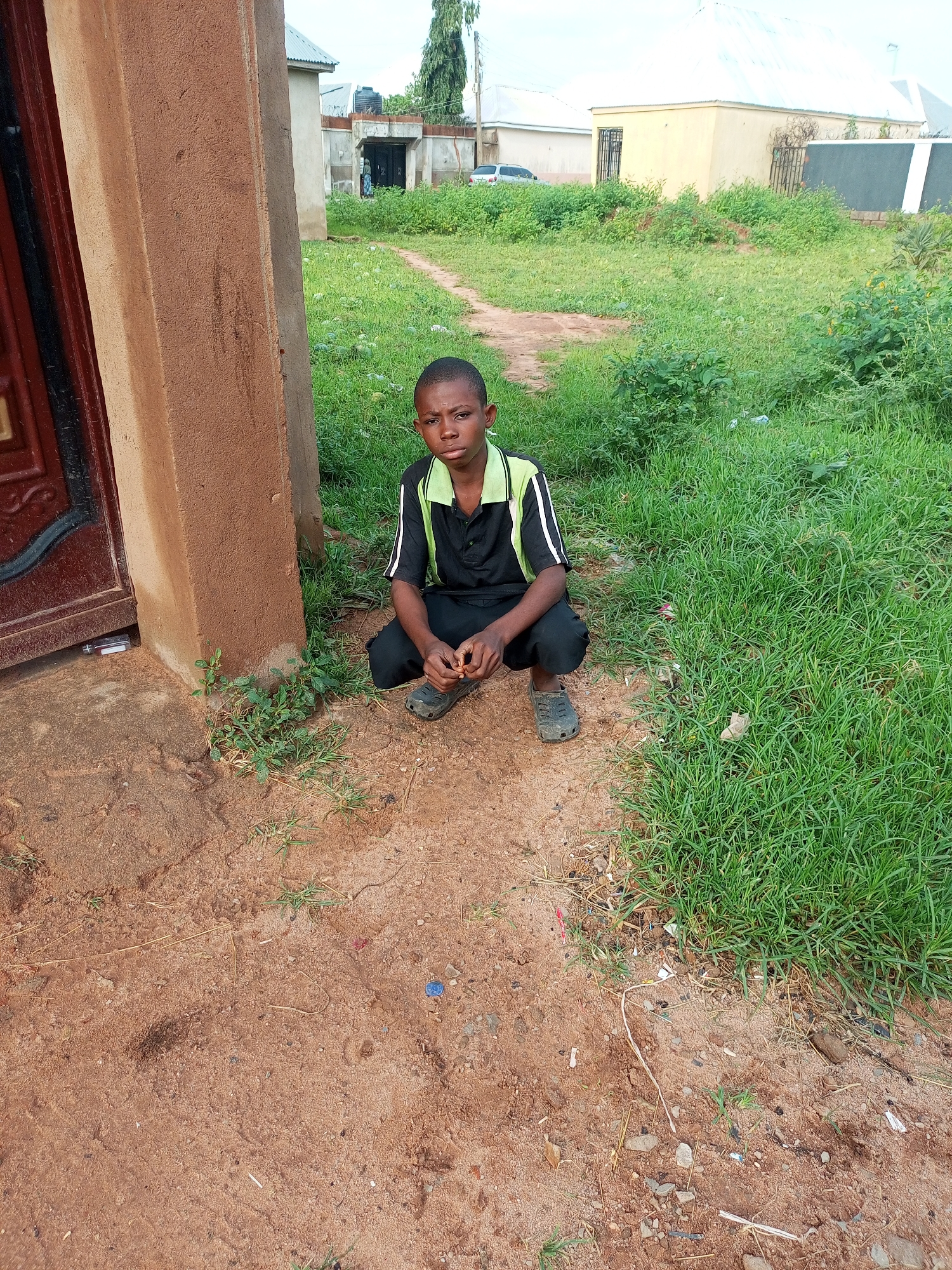 A young boy, appearing to be of African descent, is crouched on sandy ground next to a brown, stucco-walled building with a dark red door. He wears a black polo shirt with green and white accents on the collar and sleeves, and black pants. His feet are clad in grey slip-on shoes. In front of him, to his right, is a patch of green grass. Behind him, the grassy area extends, with a dirt path winding through it. Several buildings are visible in the background, including a yellow building with barred windows and a white-roofed structure. The sky is overcast. The scene is set in the city of Tempure, Nigeria.