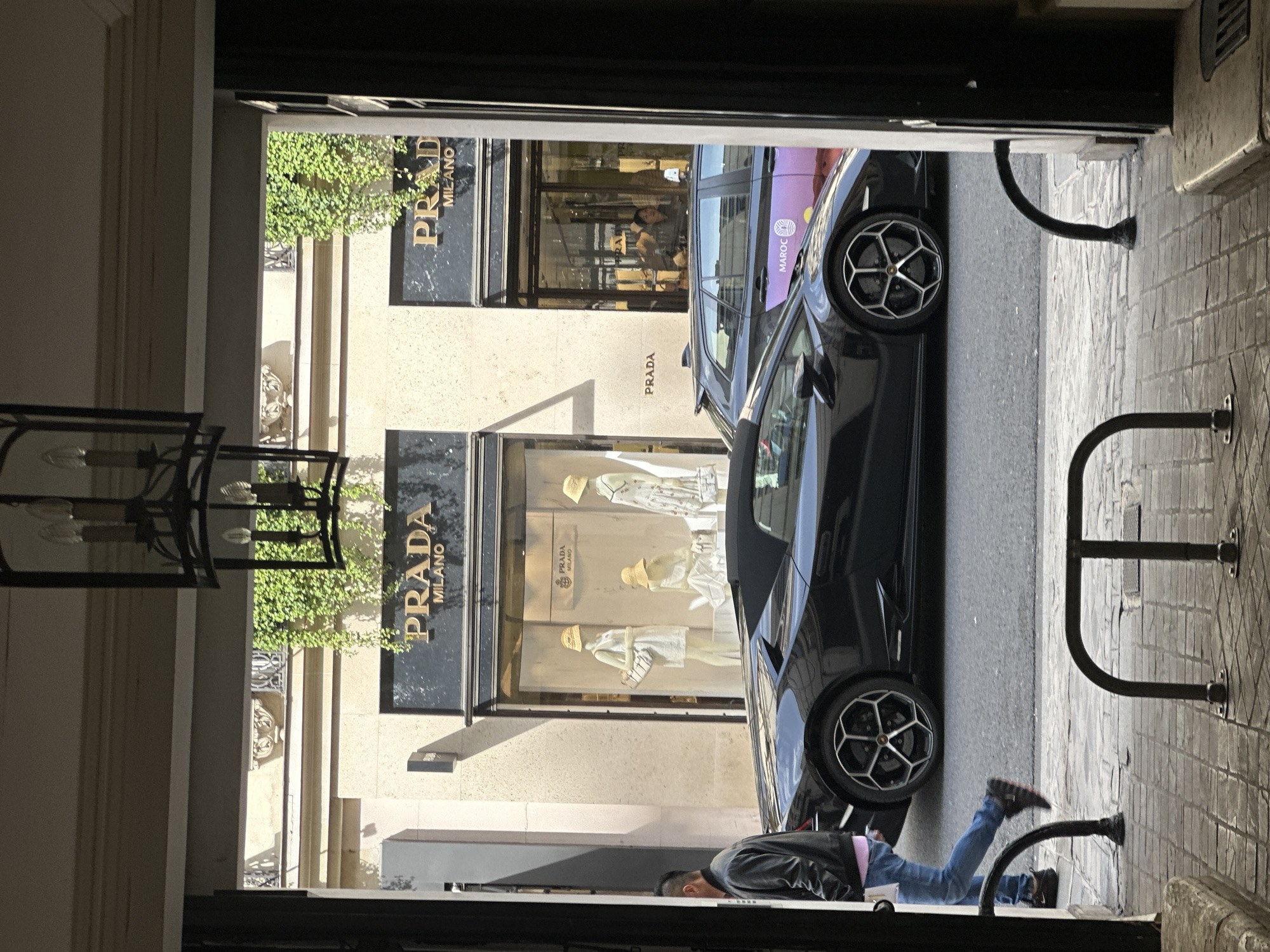 The image depicts a street scene in Paris, France. A black luxury car, possibly a Lamborghini, is parked on the street in front of a Prada store. A man wearing a leather jacket and jeans is lying on the ground near the car. The photo is taken from a doorway, showing a partial view of a light fixture and metal barriers. The Prada store's window displays mannequins with hats and handbags. A building with ornate architectural details and greenery is visible above the Prada store.