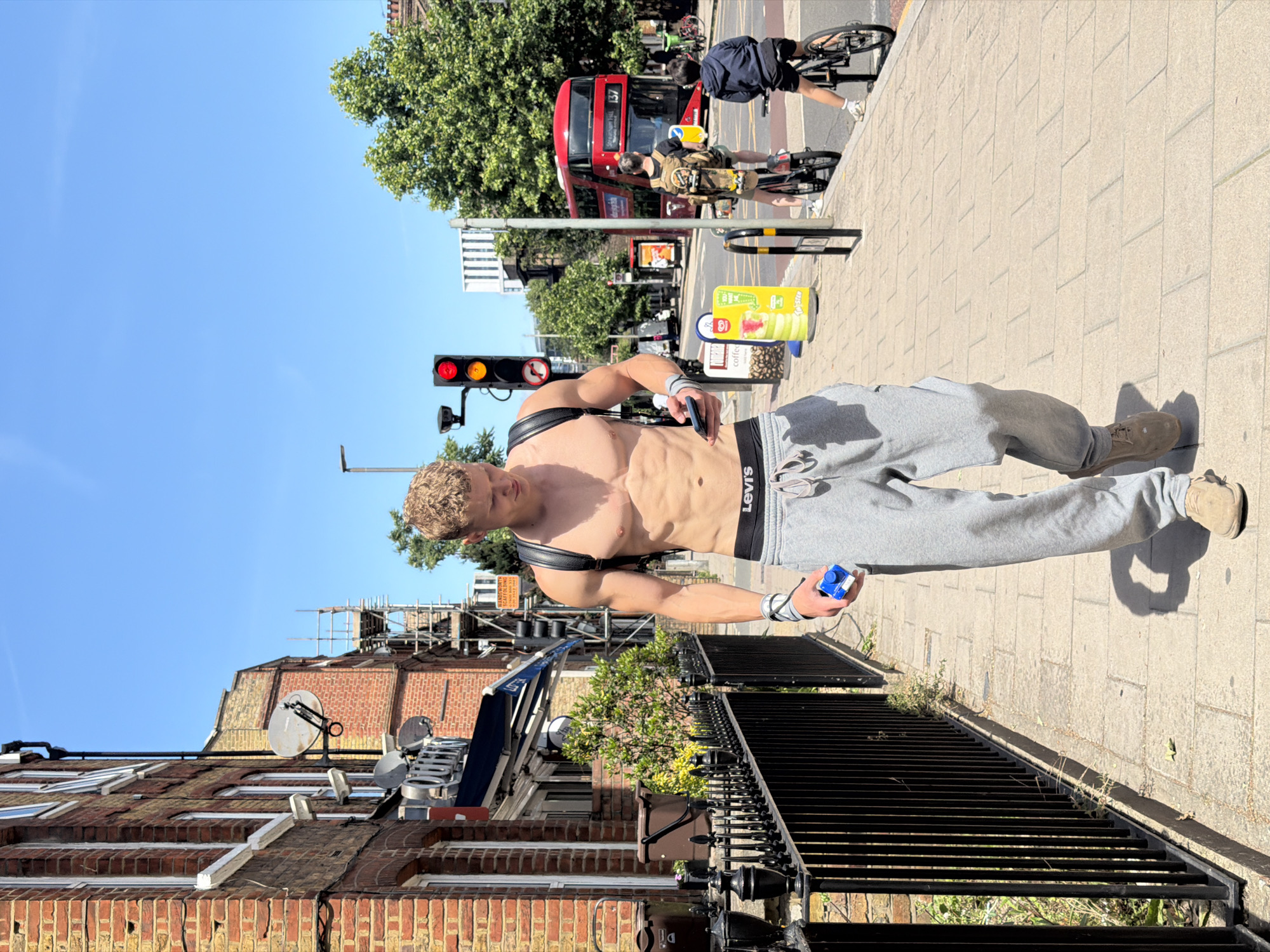 A shirtless man with blonde hair walks down a sidewalk in London, United Kingdom, on a sunny day. He is wearing grey sweatpants with "Levi's" branded underwear peeking out, and brown suede shoes. He carries a black backpack and holds a blue object in his right hand, while looking at his phone in his left hand. The sidewalk is made of light-colored paving stones and is bordered by a black metal fence. Behind the fence, a brick building with white-framed windows and satellite dishes is visible. In the background, a red double-decker bus with the number "137" displayed on its front is driving on the street. Other individuals on bicycles are also present in the background. Traffic lights and trees line the street, suggesting an urban environment.