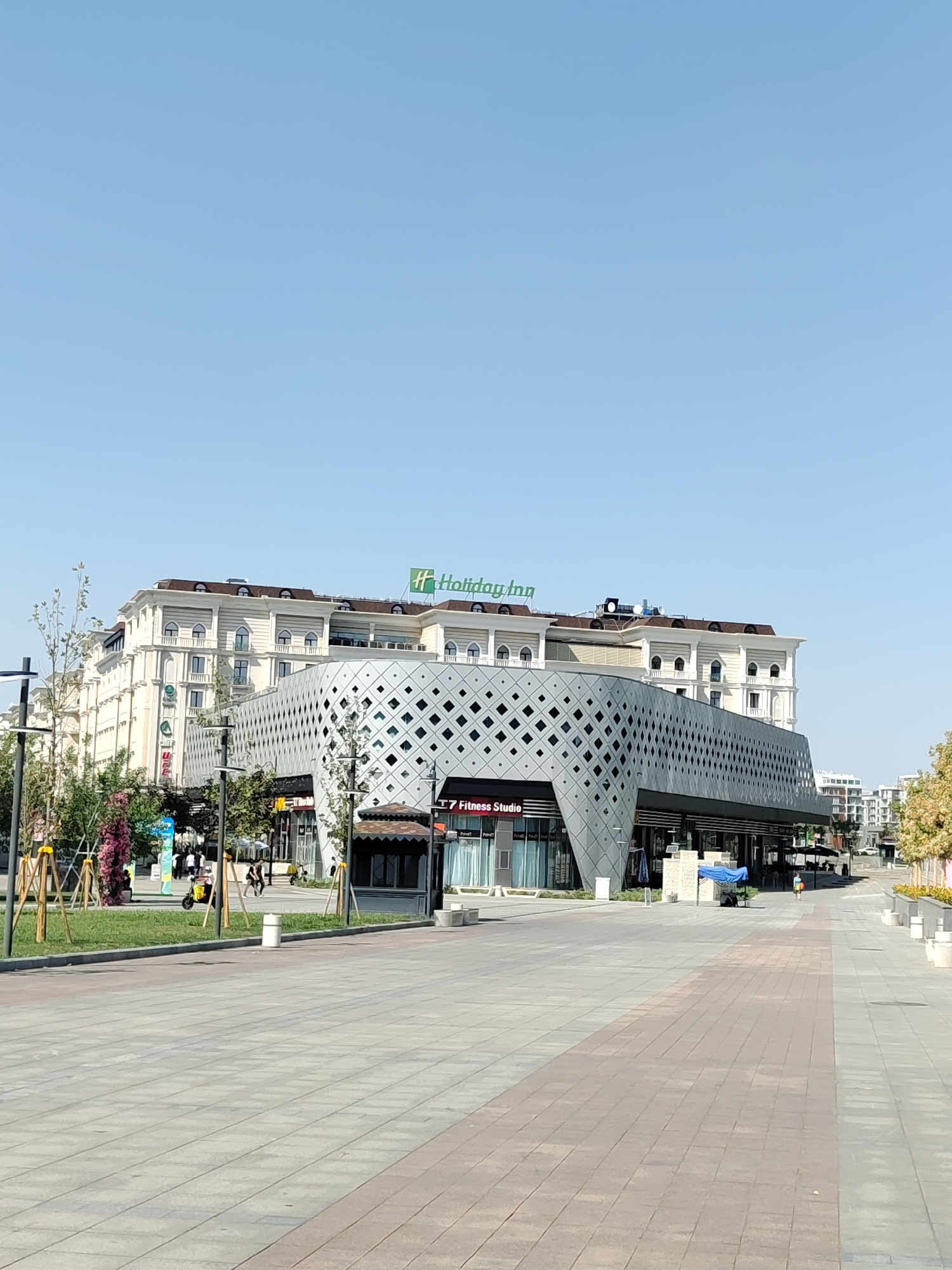 A wide, paved walkway with a clear blue sky overhead leads towards a modern building complex in Tashkent, Uzbekistan. The building features a striking, geometric facade of interlocking diamond shapes in a muted gray, which contrasts with the more traditional, ornate architecture of the hotel above it. The hotel, identified by a green "Holiday Inn" logo, has a light-colored, classical facade with arched windows and a dark-tiled roof.
On the left side of the walkway, there is a grassy area with trees and some decorative pink flowers. Several lampposts and decorative wooden structures are also visible in this area. A few people are walking along the path, and a yellow electric scooter is parked near the grassy section. On the right side of the walkway, there are storefronts with awnings and seating areas. One storefront prominently displays "T7 Fitness Studio" in a red and white sign. Further down the walkway, more buildings and trees can be seen, suggesting a commercial or residential district. The overall scene depicts a well-maintained urban environment on a clear day.