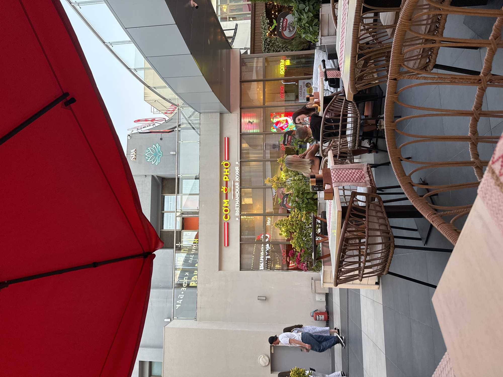 This is a ground-level, eye-level, outdoor shot of a cafe terrace in Sochi, Russia, under a large red umbrella. In the background, the facade of a building with signage for "COM PHO" and "ВЬЕТНАМСКАЯ КУХНЯ" (Vietnamese Cuisine) is visible. A group of people are seated at tables, with two women and a child at one table, and another woman at a separate table. Tables and chairs are arranged on the terrace. The flooring is a mix of dark gray and light gray tiles. A fire extinguisher is visible near a door on the left side of the image. The overall ambiance suggests a casual dining environment on a pleasant day.