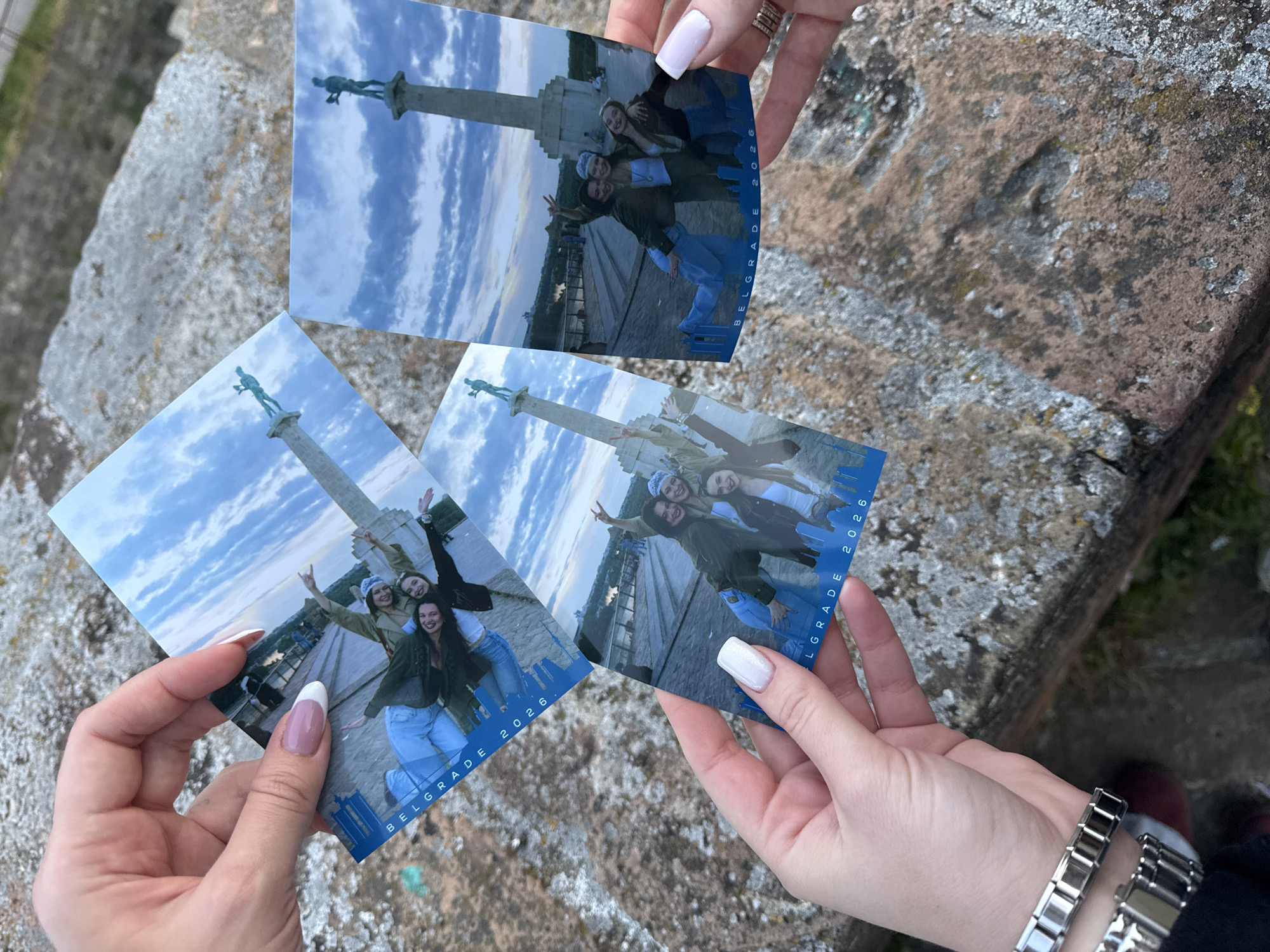 Three photographs are held by two sets of hands, seemingly outdoors. The photographs feature a monument with a statue atop a pillar against a cloudy sky, and a group of four young women posing together in front of the monument. The photographs have a blue border with "BELGRADE 2026" written on it. The hands holding the photos have manicured nails. One hand has a silver watch. The photos are resting on a stone or concrete surface. Based on the monument shown in the photographs, the location is likely the Victor Monument in Belgrade, Serbia.