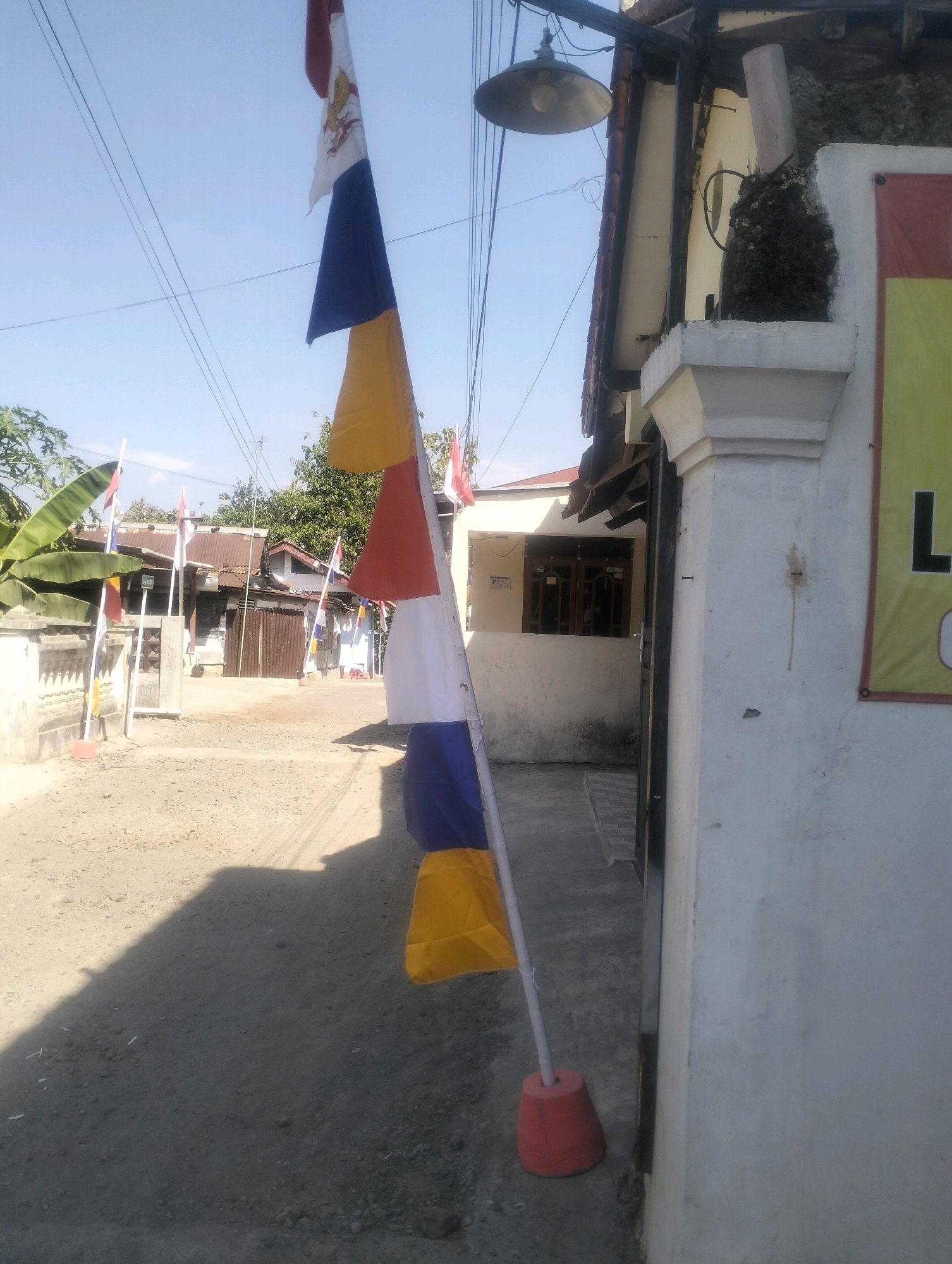 A street-level, eye-level, medium shot captures a section of a residential street in Purwokerto, Indonesia, under a clear blue sky. In the foreground, a tall, thin flagpole stands vertically, adorned with a series of horizontally striped flags in red, white, blue, and yellow, with the topmost flag also featuring a white emblem. The flagpole is anchored by a conical, red base resting on the dusty, unpaved road. To the right, a white-painted pillar and a section of a building wall are visible, with a partial banner displaying the black letter "L" on a yellow background. A rustic light fixture is attached to an overhang above.
The background reveals a street lined with modest, single-story houses with corrugated metal roofs and simple facades. Some houses have visible windows and doors, and one appears to have a gate. Small, decorative flags on shorter poles are also placed along the fence line of one of the properties. Trees and foliage are visible behind the houses, and utility lines stretch across the sky. The overall impression is of a quiet, residential neighborhood in a tropical climate. The lighting suggests it is daytime, with clear visibility and shadows cast by the objects.