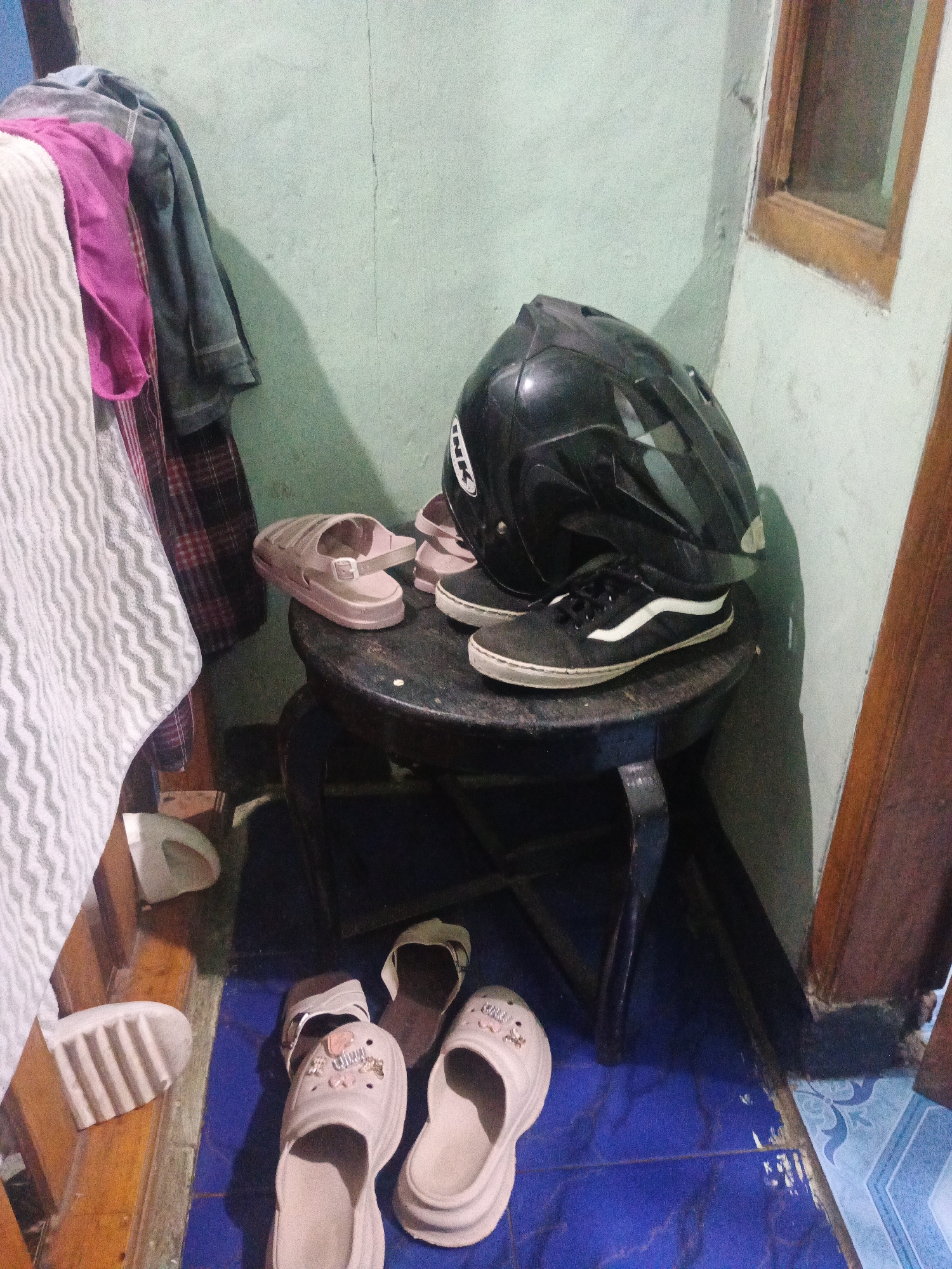 The image depicts a cluttered corner of a room, possibly a dwelling in Sumedang, Indonesia, characterized by a light green painted wall and a tiled floor of alternating blue and white patterns. On a dark, ornate wooden stool in the center of the frame, a black motorcycle helmet rests atop a pair of black Vans sneakers. To the left of the stool, a pile of clothing is draped over something not fully visible, including a pink garment and what appears to be a patterned fabric. On the floor, several pairs of sandals are scattered. Two light pink platform sandals with embellishments are prominent in the foreground, alongside a pair of dark brown slides. A white sandal with ribbed detailing lies near the bottom left of the image. The lighting is dim, casting shadows that contribute to the overall sense of disarray. The wooden doorframe on the right side of the image appears aged and slightly worn.