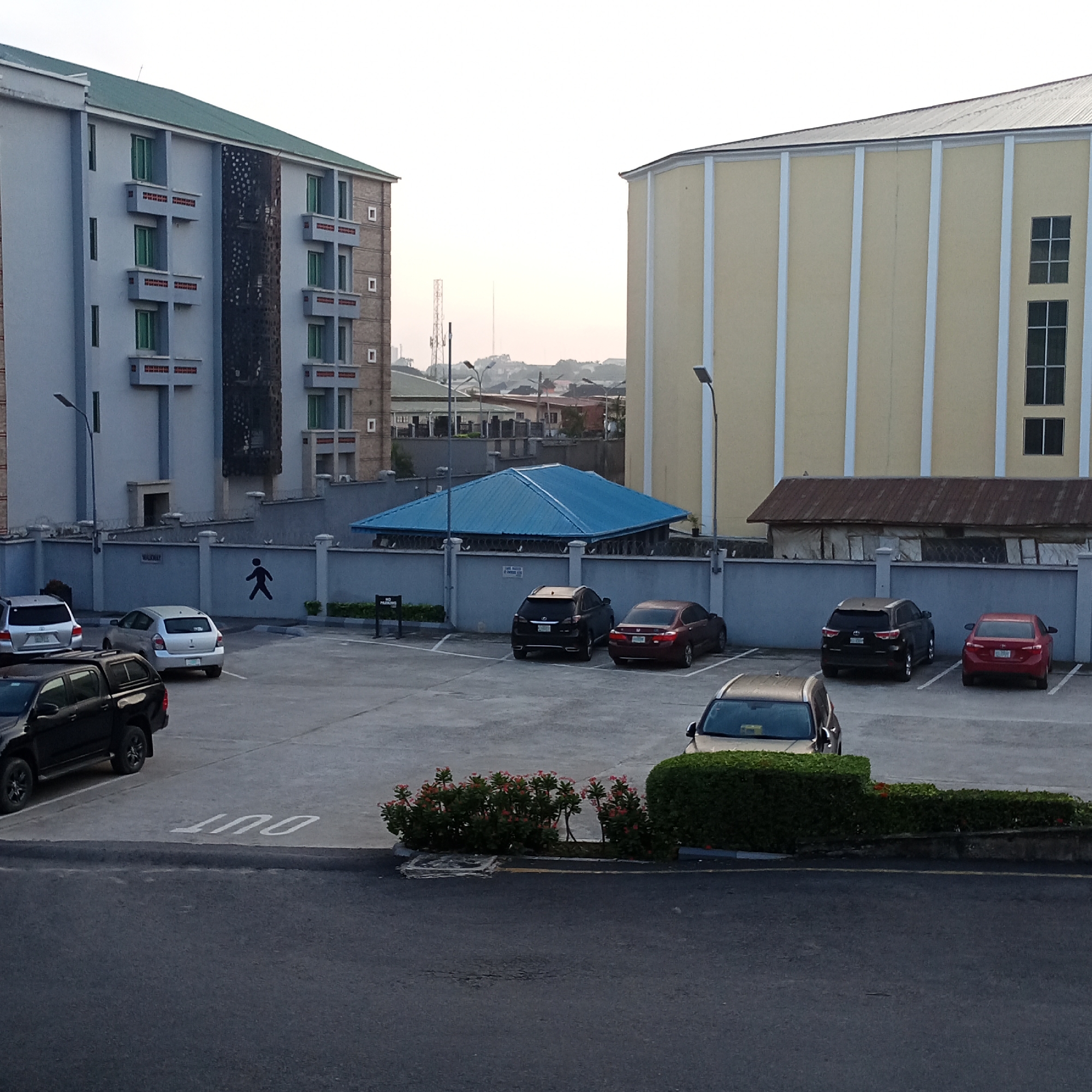 The image shows a parking lot outside two buildings in Ibadan, Nigeria. The building on the left is a light blue, multi-story building with green-roofed balconies. The building on the right is a pale yellow, multi-story building with white vertical trim. A blue-roofed structure is visible between the two buildings. Several cars are parked in the lot, including a black SUV, a silver sedan, a black SUV, a red sedan, and a silver SUV. A sign with a silhouette of a person walking is painted on a gray wall. The ground is paved with asphalt and concrete. There are some small bushes and flowers planted in the parking lot. The sky is overcast.