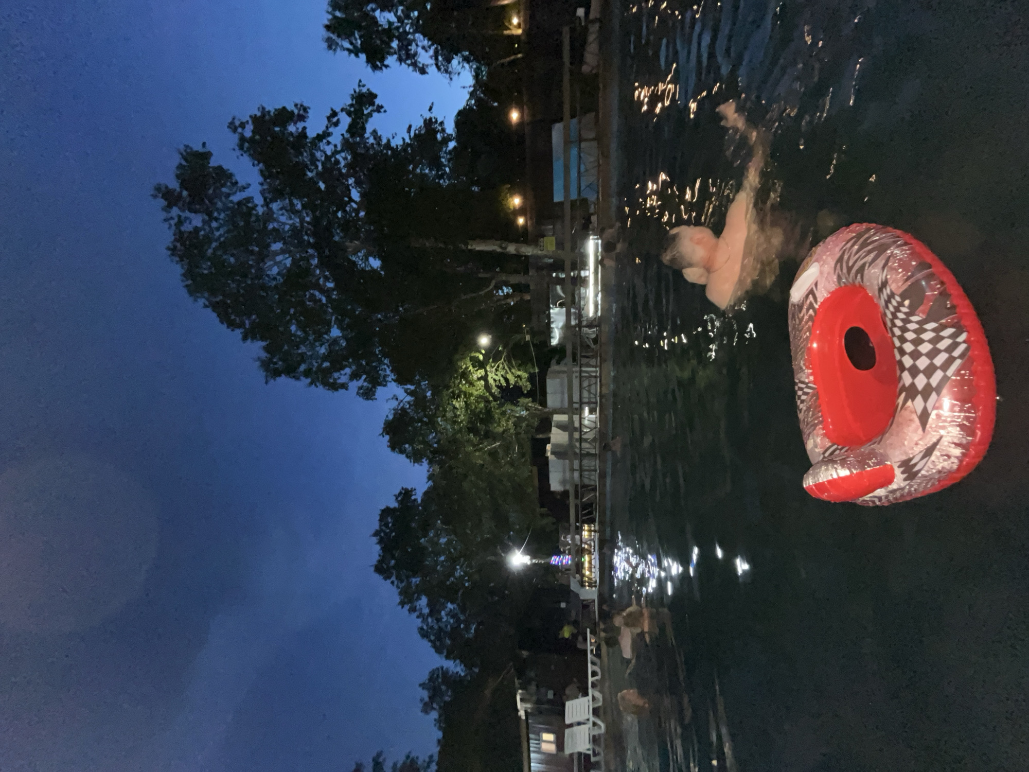 At dusk in Paratunka, Russia, people relax in a dark, outdoor swimming pool illuminated by reflections of lights on the water's surface and a string of colorful lights on a pole. An inflatable ring with a checkered pattern and red inner rim floats in the foreground. In the background, trees and a wooden structure are visible, with lounge chairs set up on the pool deck. The sky is a deep twilight blue.