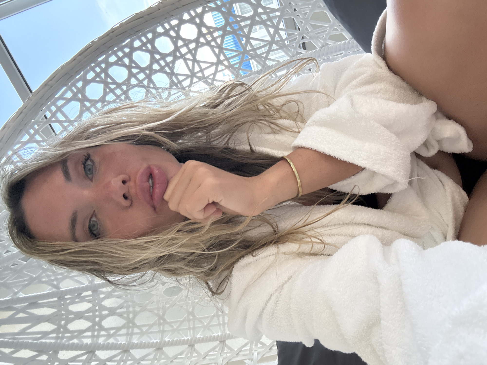 A woman is photographed from a low angle, lying down. She is wearing a white bathrobe. Her long, blonde hair is wet and falls around her face. She has green eyes and is looking at the camera. Her lips are slightly parted, and her right hand is resting on her chin. She is wearing a thin gold bracelet on her right wrist. The background is a white wicker chair with a blue sky visible through it. The overall lighting suggests it is daytime. The image is oriented vertically.