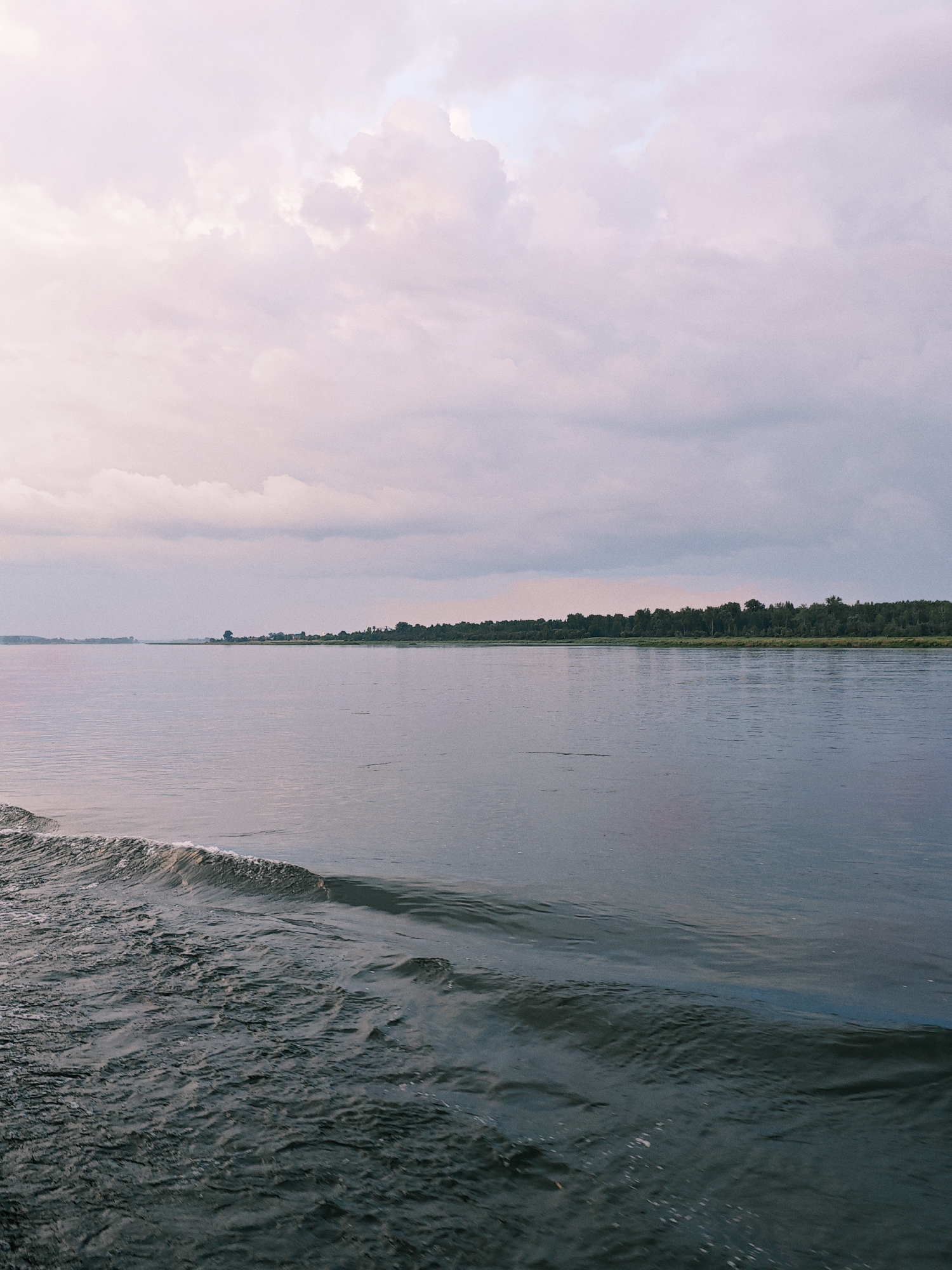 The image depicts a wide, calm river in Kazachinskoe, Russia, under a cloudy sky. The water is a deep blue-gray, with a gentle wave breaking on the left side of the frame, indicating movement or passage. The river stretches towards a distant, tree-lined shore, where a thin strip of green foliage separates the water from the sky. In the background, a few structures are visible along the shoreline, suggesting a small settlement. The sky is filled with soft, diffused clouds, tinged with pink and pale purple hues, creating a serene, atmospheric backdrop. No people or specific actions are identifiable in the image.