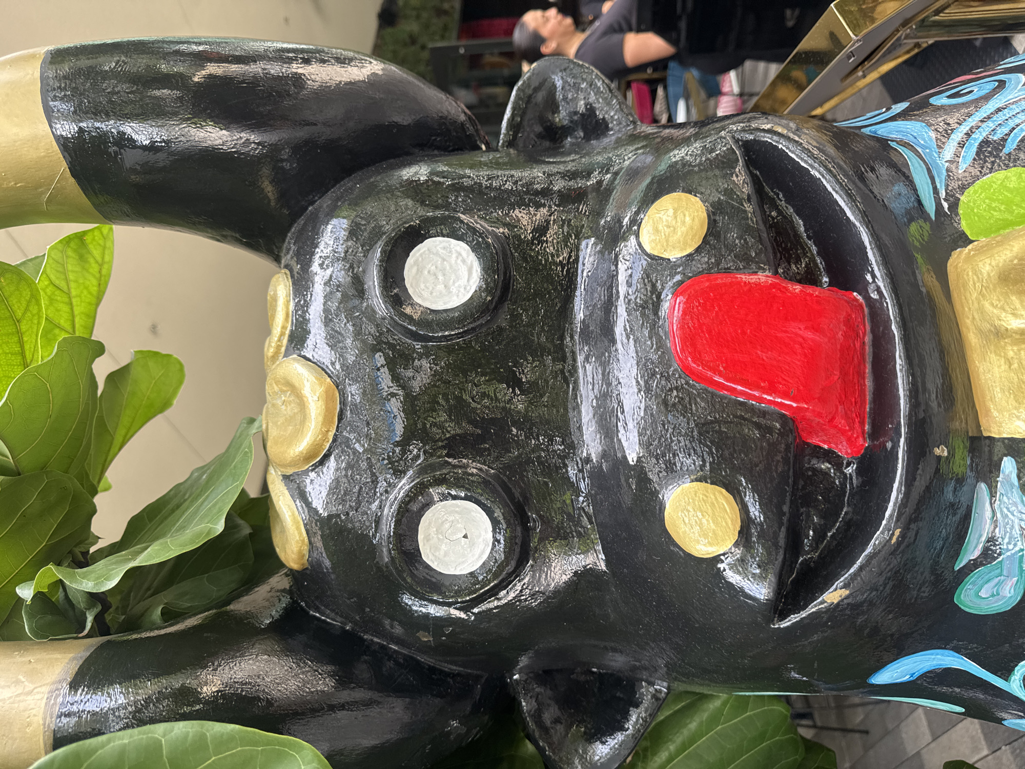 In Aventura, Florida, a close-up, eye-level, horizontal shot captures the head of a black, glossy sculpture resembling a stylized bull or cow. The sculpture features large, circular white eyes, gold accents around the eyes and forehead, and a bright red tongue sticking out. The sculpture is adorned with blue and green painted swirls on its lower body, which is partially visible. Lush green foliage, likely from a Fiddle Leaf Fig plant, is present behind and to the left of the sculpture, obscuring parts of the background. In the blurred background to the right, a woman with dark hair pulled back is visible, wearing a black top and standing near a reflective gold railing. The setting appears to be outdoors, possibly a public space or entrance area with a paved walkway.