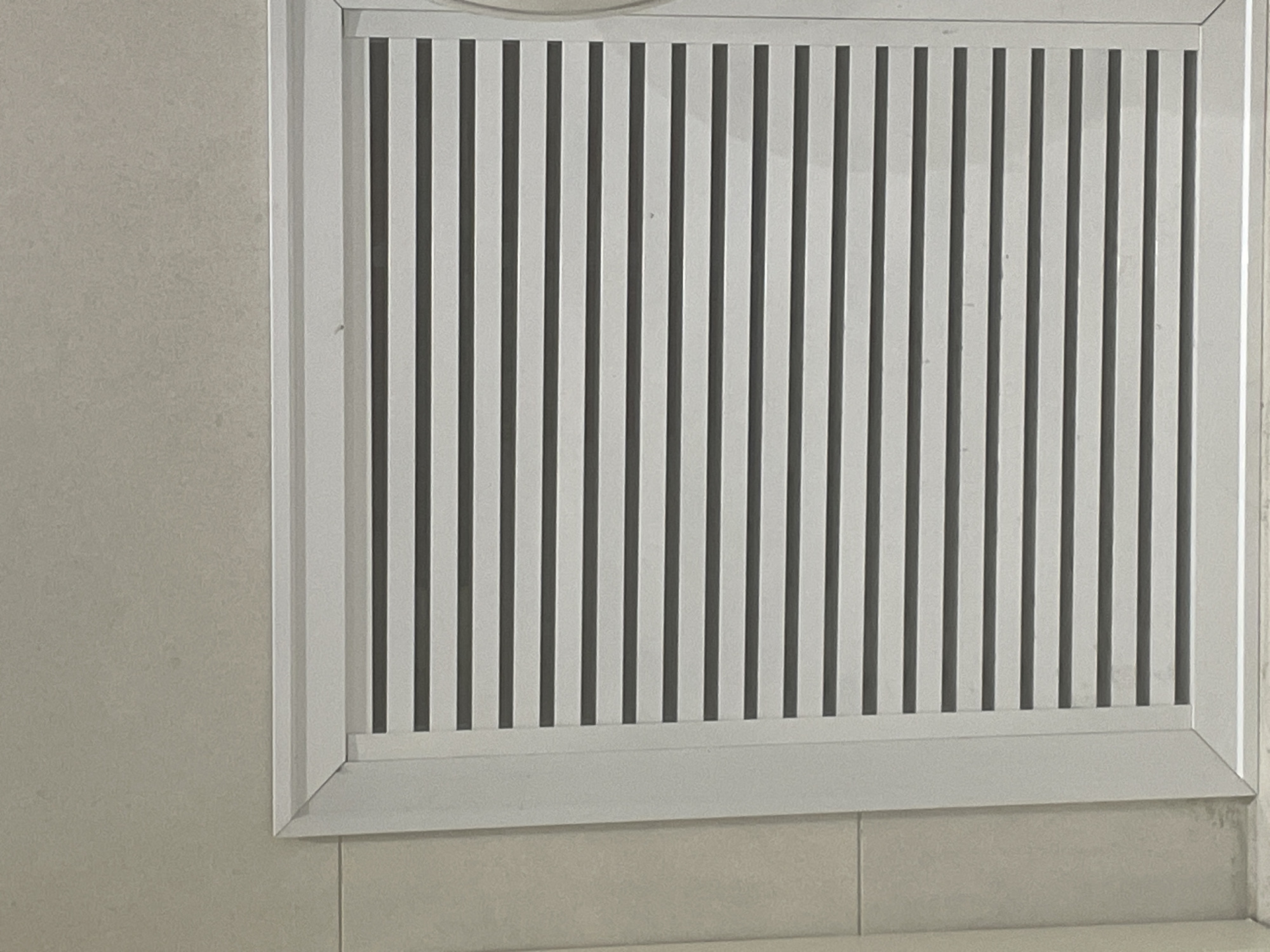 The image shows a close-up of a white louvered vent set into a beige tiled wall. The vent is rectangular with horizontal slats. The wall is tiled with large, square, beige tiles. The lighting appears to be artificial and overhead, casting a soft, even light on the scene. The image is taken from a medium distance, with the vent filling most of the frame. The overall impression is one of a clean and functional indoor space. There is no indication of the city of Yunus Emre, Turkey, in the image itself.