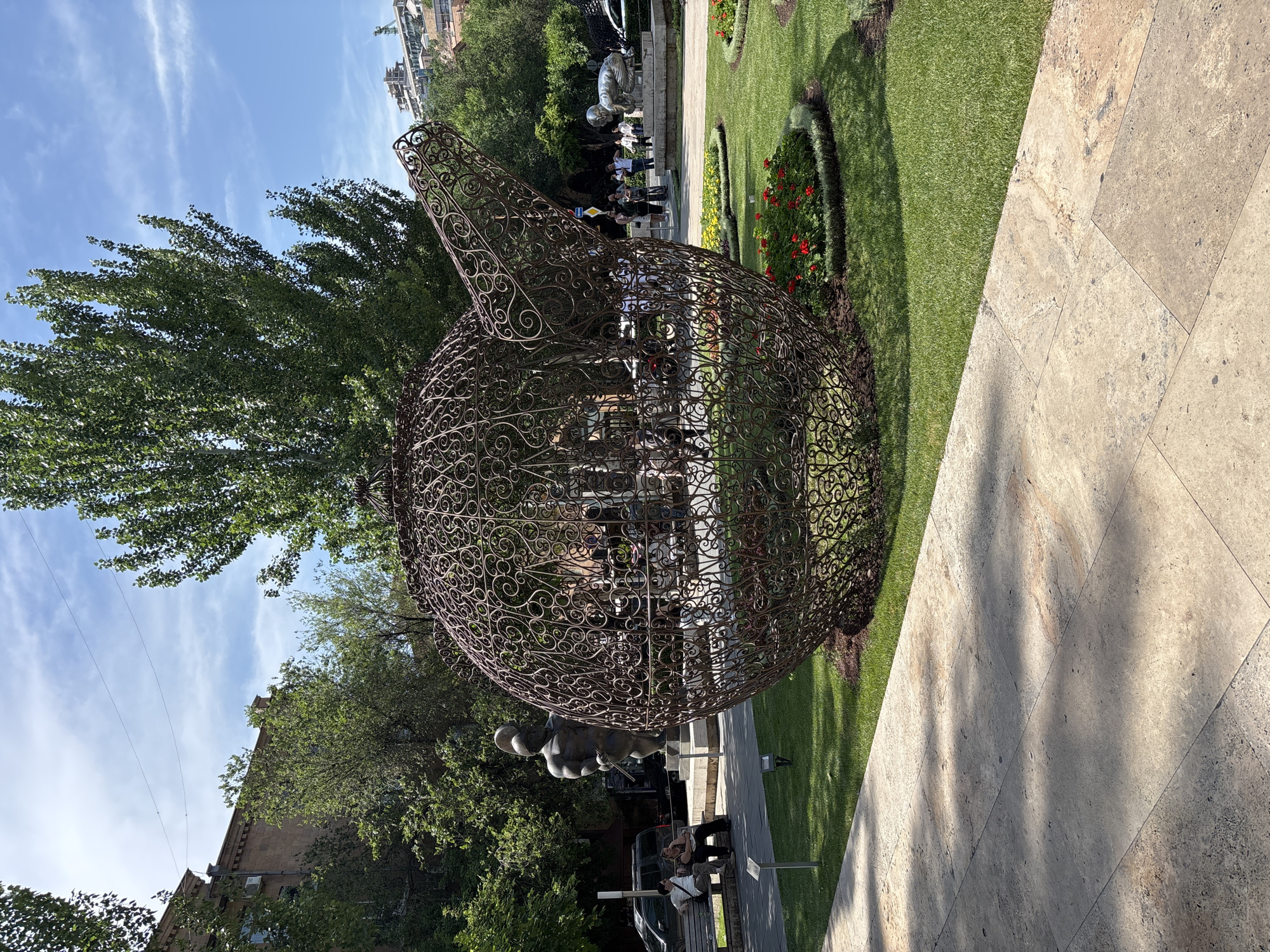 The image depicts a large, ornate wire sculpture of a teapot in a park setting. The sculpture is made of intricate metalwork, forming swirling patterns. It is situated on a manicured lawn with circular flower beds containing red and yellow flowers. In the background, there are other sculptures, including a statue of a muscular male figure and a smaller sculpture of a crouching child. Several people are visible in the background, some walking and others sitting on a bench. A paved walkway runs in the foreground, and trees and buildings are visible beyond the park. The scene is set under a clear blue sky with scattered clouds. This is the Cascade complex in Yerevan, Armenia, a giant stairway adorned with sculptures and fountains.