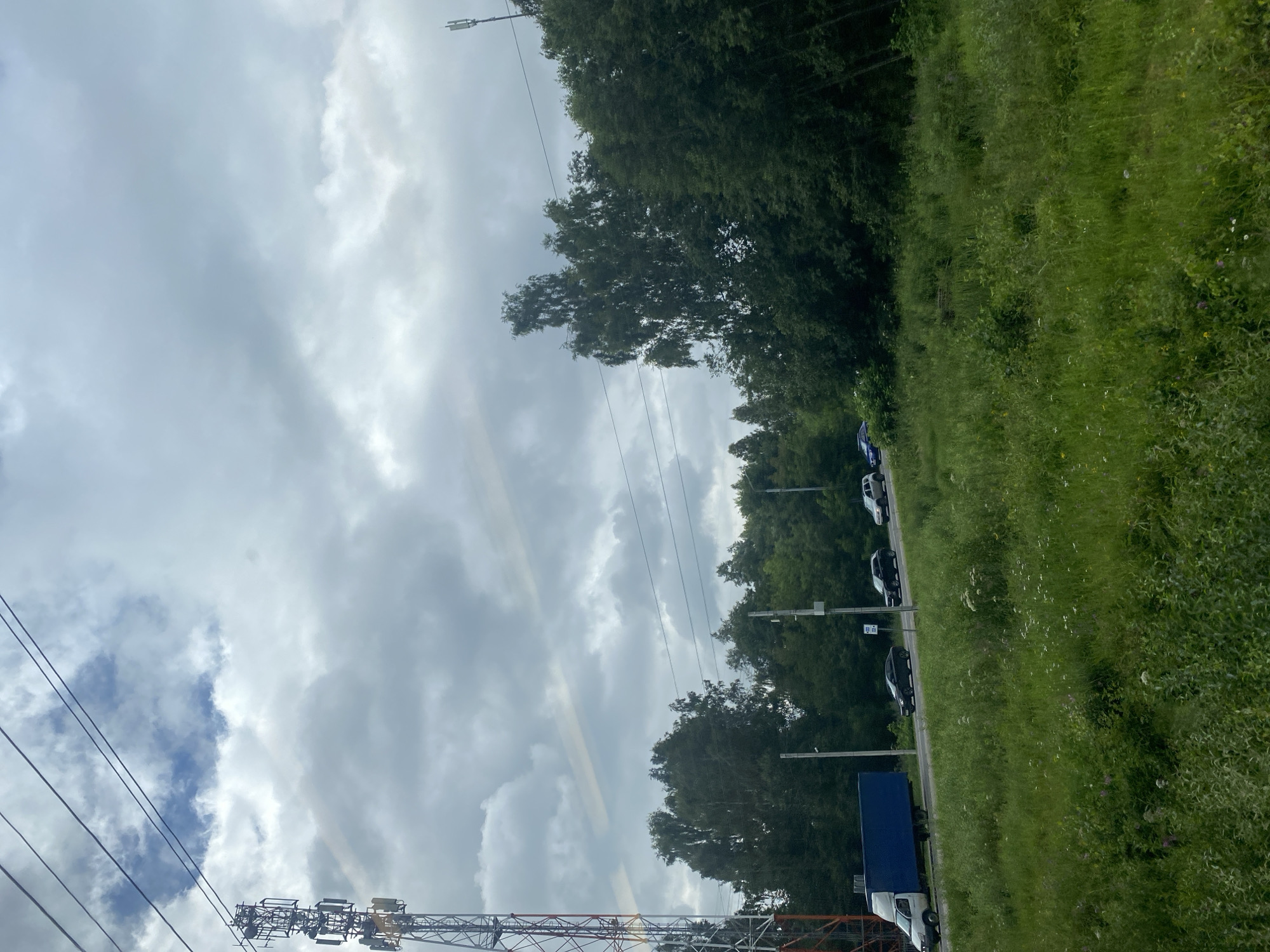 An outdoor, eye-level, slightly tilted, medium shot captures a roadside scene on a cloudy day. In the foreground, tall green grass and scattered white wildflowers line a slight incline. A paved road runs horizontally across the midground, with a line of trees and dense foliage forming a backdrop. A white truck with a blue cargo box is traveling on the road from left to right, followed by several passenger cars. A tall, lattice radio tower with multiple antennas stands on the left side of the image. Power lines traverse the sky, intersecting with the top of the radio tower. The sky is filled with thick, grey clouds, with hints of blue peeking through. The environment suggests a location on the outskirts of an urban area, possibly near a highway. The scene is devoid of any visible people. The image appears to have been taken in Dubna, Russia, a city known for its scientific research institutions and its proximity to the Volga River, often characterized by such roadside infrastructure and natural landscapes.