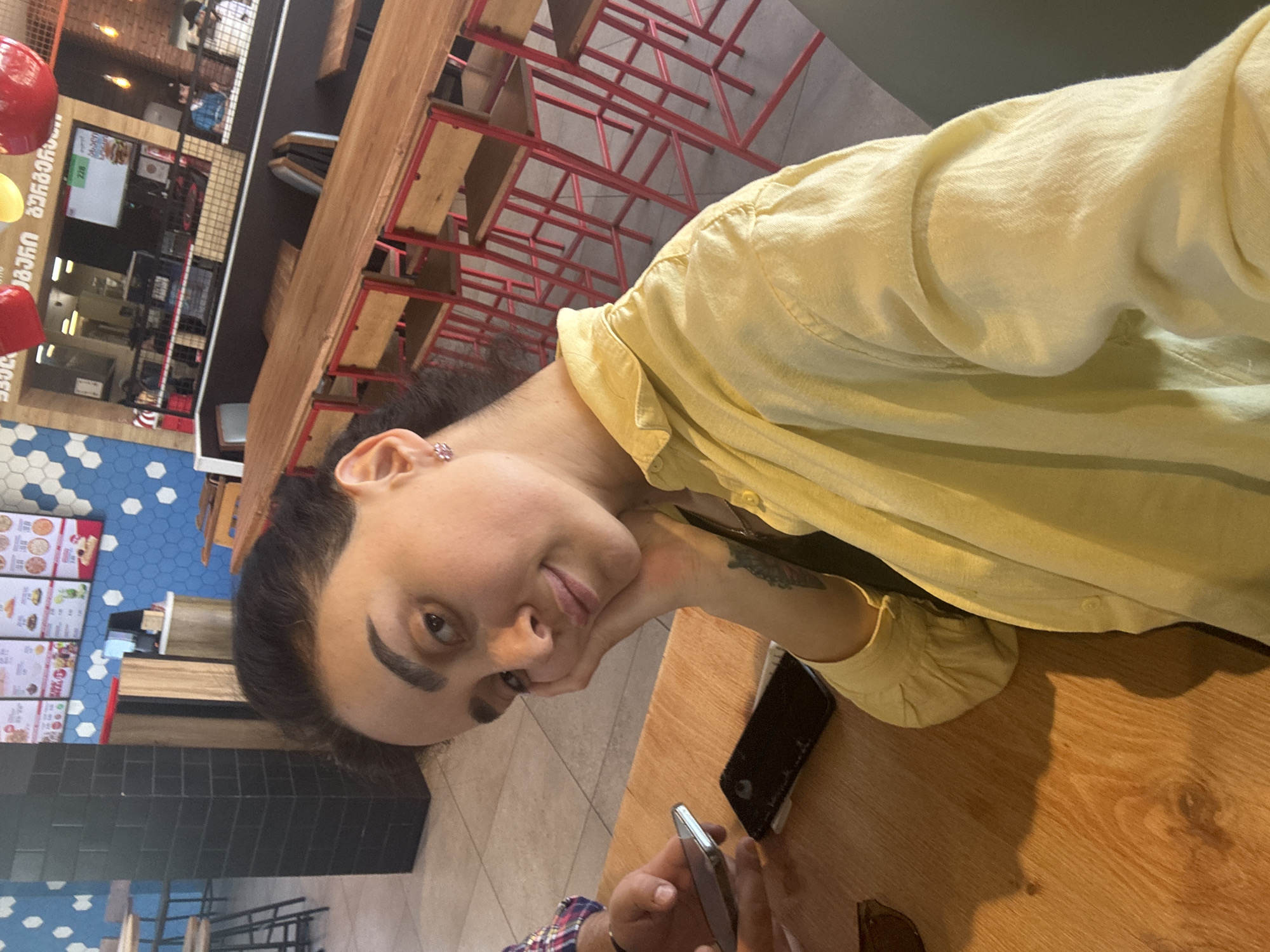 The image shows a person in a light yellow shirt at a table in a fast-food restaurant. A black smartphone is on the table next to them. Another person’s hands holding a silver smartphone are visible at the bottom of the frame. The restaurant features red metal chairs and wooden tables. In the background, there is a menu board written in Georgian, and blue and white tiled walls. The location is identified as Tbilisi, Georgia based on the Georgian language.
