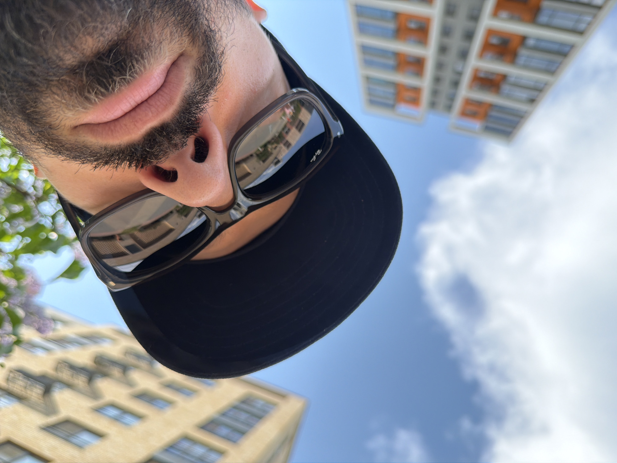 The image is a low-angle, upside-down selfie. The individual has a dark beard and mustache, fair skin, and is wearing dark sunglasses and a black baseball cap. The sky is visible in the background, with scattered clouds. Two buildings are visible, one with a light yellow exterior and rows of windows, and another taller building with a white facade and orange accents. The presence of buildings suggests an urban setting. The specific location in Tyumen, Russia, cannot be determined with the limited information provided in this image.