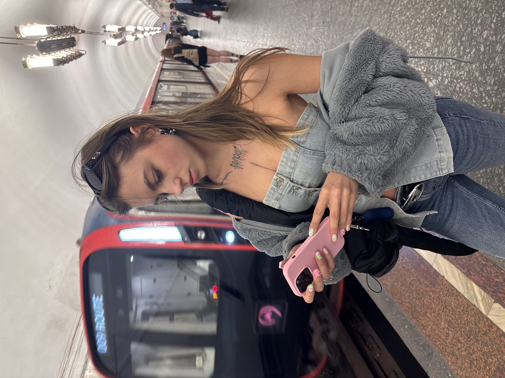 A young woman with long, light brown hair stands on a subway platform in Moscow, Russia. She is wearing a denim tube top, a grey fuzzy jacket draped over her shoulders, and blue jeans. She is looking down at her pink phone, which has a decorative case. A train is approaching on the tracks behind her, its headlights illuminating the platform. Other passengers are visible in the background, waiting for the train. The platform is made of stone, and the ceiling is arched and lit by overhead lights.